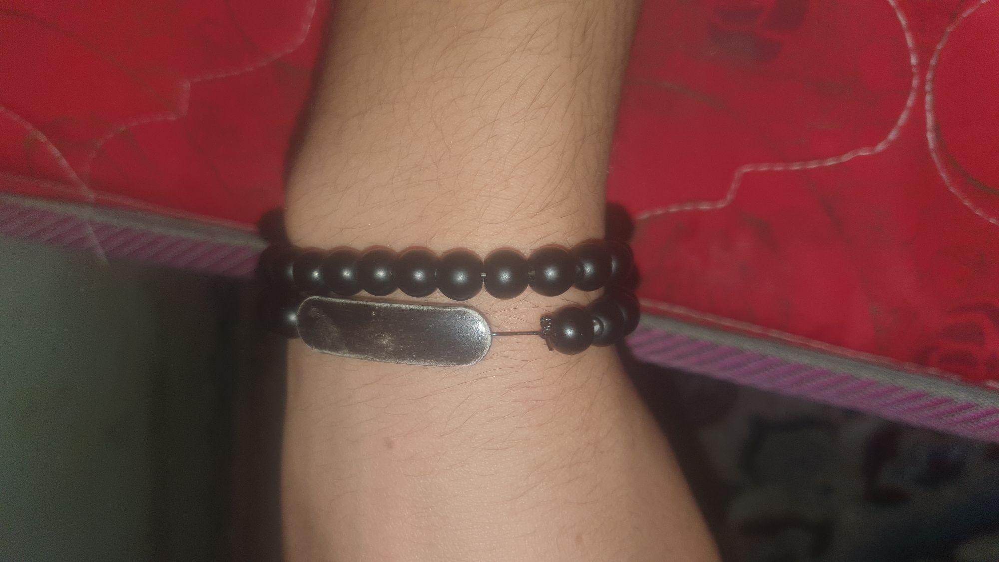 The image shows a close-up of a person's wrist, adorned with two beaded bracelets. One bracelet is composed of black, matte beads, while the other features a single, rectangular, metallic pendant with a dull finish. The pendant is attached to the black beaded bracelet by a thin black string. The wrist is positioned against a textured surface, appearing to be a red fabric with a subtle pattern. The background is dark and out of focus, with hints of purple and white stripes visible on the left side. The scene appears to be indoors, and the lighting suggests it may be dimly lit. There is no discernible urban environment or specific location within a city.