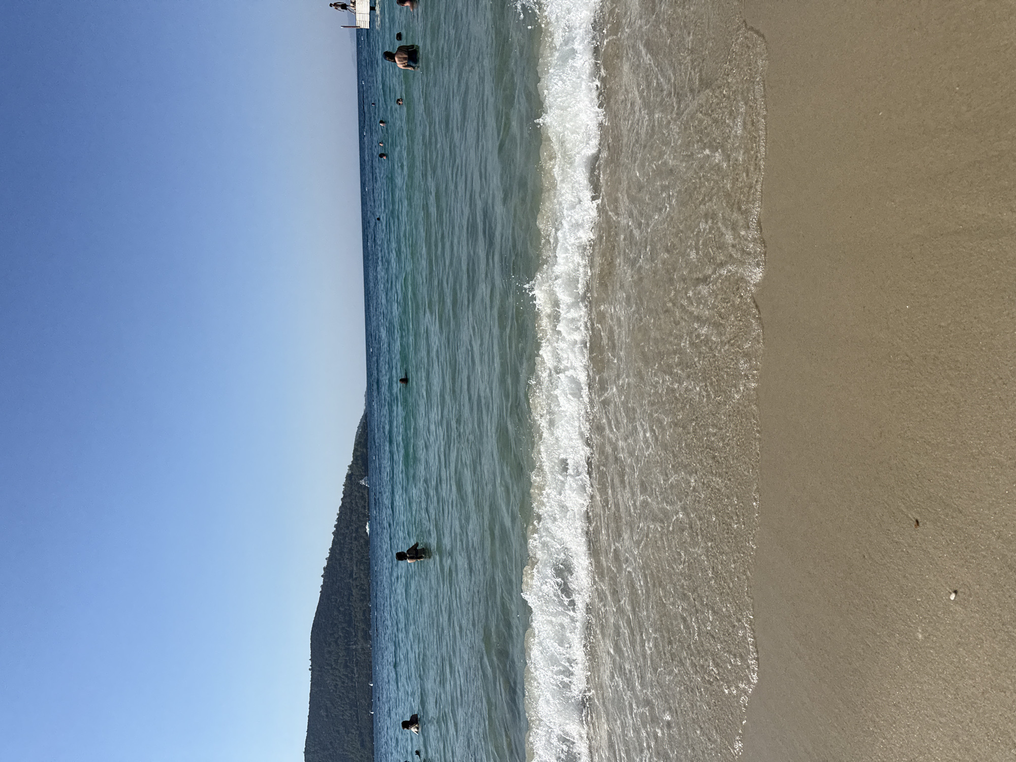 The image depicts a serene beach scene in Bodrum, Turkey. The foreground is dominated by the wet sand of the beach, with a gentle wave lapping at the shore. The water is clear and turquoise, revealing the sandy bottom. Several people are visible in the water, swimming and enjoying the calm sea. In the background, a lush green island with trees rises from the water, creating a picturesque backdrop. The sky is a clear, cloudless blue, indicating a sunny day. A small white structure, possibly a pier or platform, is visible on the right side of the image, with a person standing on it. Several boats are also scattered in the distance. The overall impression is one of a tranquil and idyllic beach vacation.