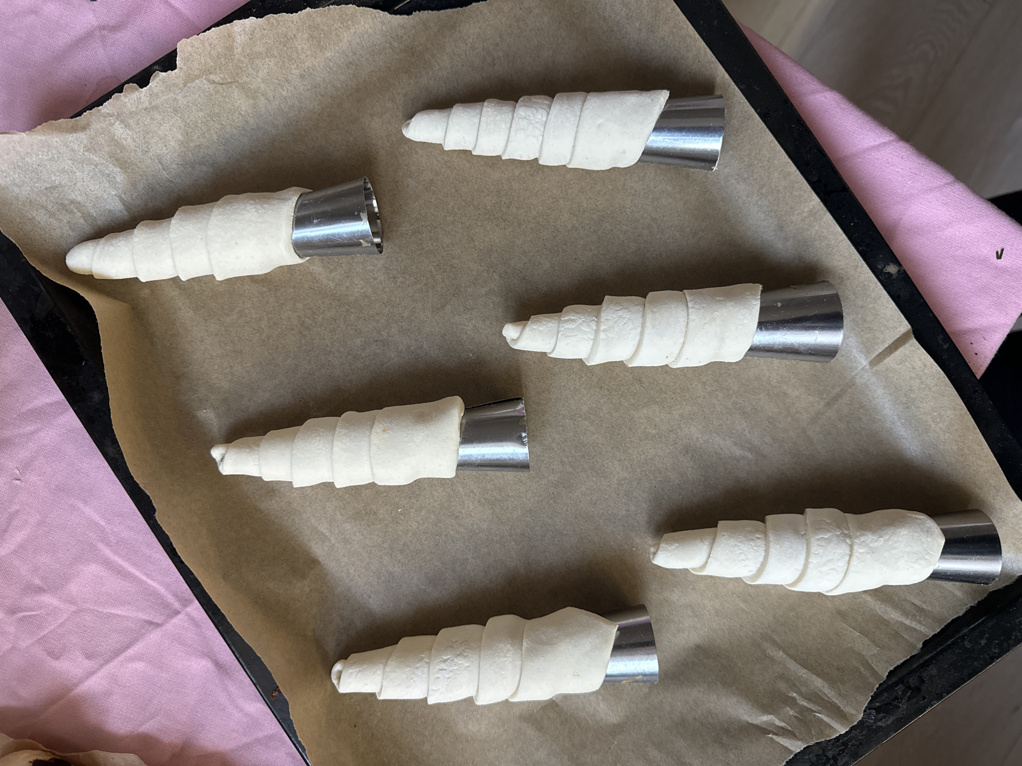 An overhead view depicts a black baking tray lined with crumpled brown parchment paper, holding six unbaked pastry items. Each item consists of a light-colored, spiral-wrapped dough cone covering a conical, silver-toned metal cream horn mold. The molds are reflective, with a wider base tapering to a rounded point, around which the dough is tightly wound. The items are irregularly spaced across the parchment paper. A segment of pink fabric is visible beneath the upper-right and lower-left edges of the baking tray, extending beyond its boundaries. Below the pink fabric, to the bottom right, a light-colored floor or surface is partially visible. The scene is located within an indoor environment in the city of Krasnoyarsk, Russia. No individuals are present.