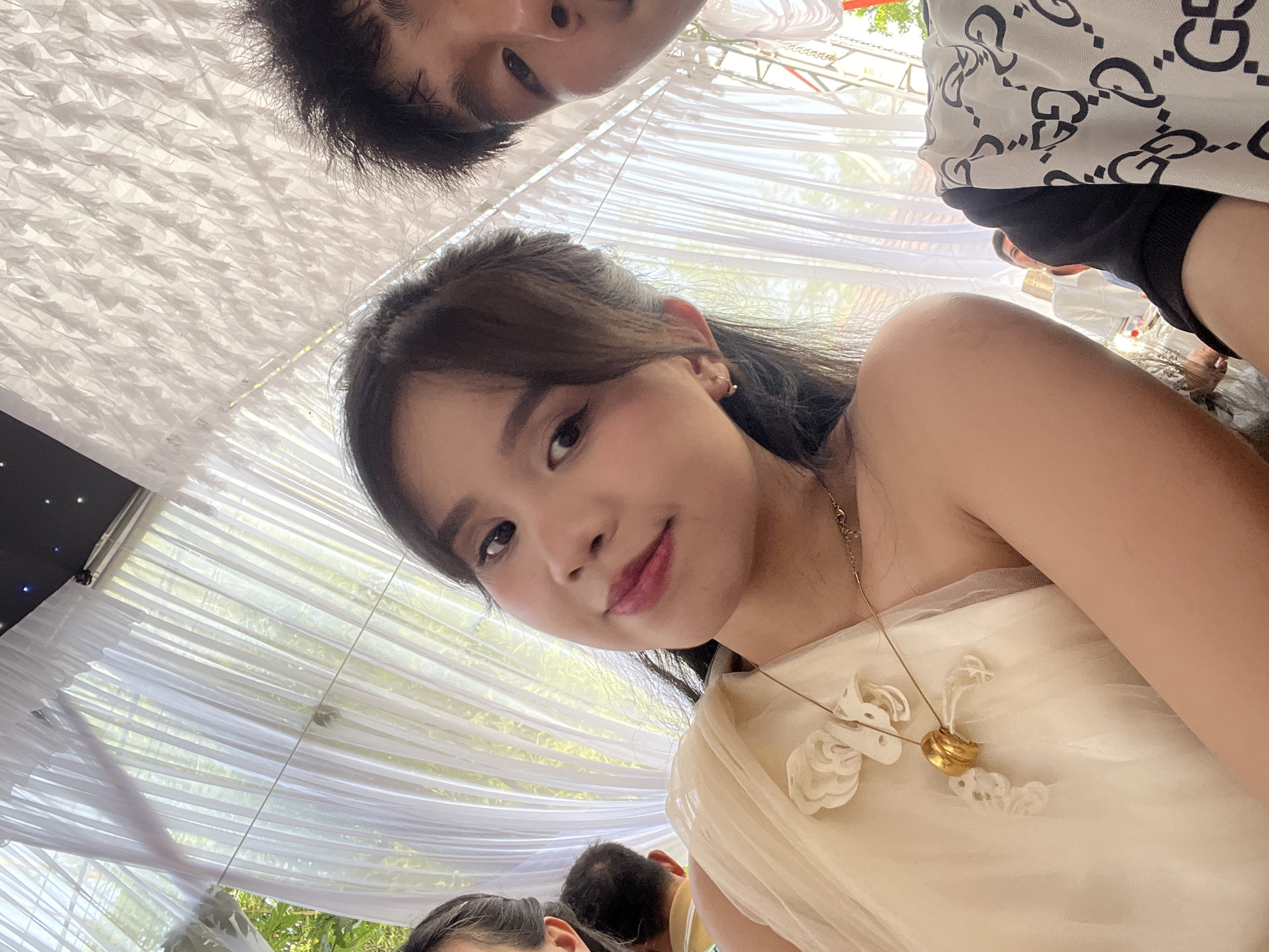This image captures a close-up, upward-angled perspective of two individuals and their surroundings, likely a selfie taken from below. The primary subject is a young woman with dark hair, looking directly at the camera with a gentle smile. She is wearing a light-colored, possibly cream or pale yellow, strapless or off-the-shoulder dress, featuring delicate floral or ruffled appliques on the bodice. A fine gold chain necklace with a prominent, round gold pendant rests on her collarbone.
Above and slightly behind her, the upper part of a man's face – including his forehead, dark hair, and eyes – is visible. He is wearing a white shirt with a repeating black "G G" pattern, resembling a well-known designer logo.
The setting appears to be an outdoor or semi-outdoor event, with a canopy of draped white sheer fabric creating a ceiling effect. Bright, diffused light filters through these drapes, indicating daytime. Green foliage and hints of natural light are visible beyond the translucent fabric, suggesting a garden or park venue. In the lower background, other blurred individuals and their heads can be seen, further suggesting a social gathering. A dark panel with small white lights is visible in the upper left corner, possibly part of a screen or decoration.
The overall atmosphere suggests a festive or celebratory event in Bình An, Vietnam, possibly a wedding or party, given the attire and decorative canopy. The direct gaze of the woman conveys engagement and a sense of personal connection to the camera.