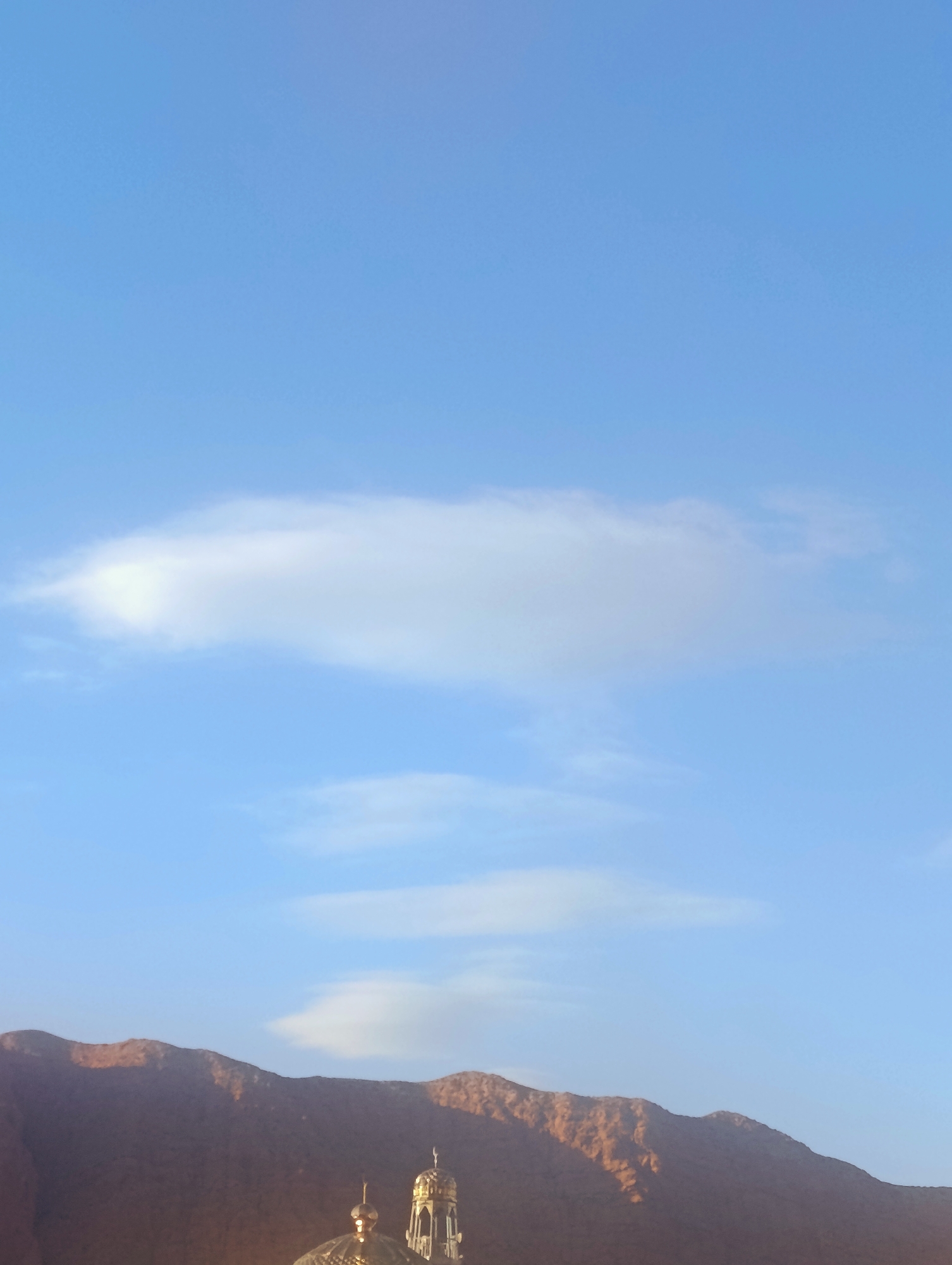 An image captures a clear, bright blue sky with scattered white clouds. In the foreground, the silhouette of a mountainous landscape is visible, with the tops of several domed structures, possibly minarets, emerging from behind the mountains. The lighting suggests it is either sunrise or sunset, as parts of the mountain are illuminated with a warm, golden hue.