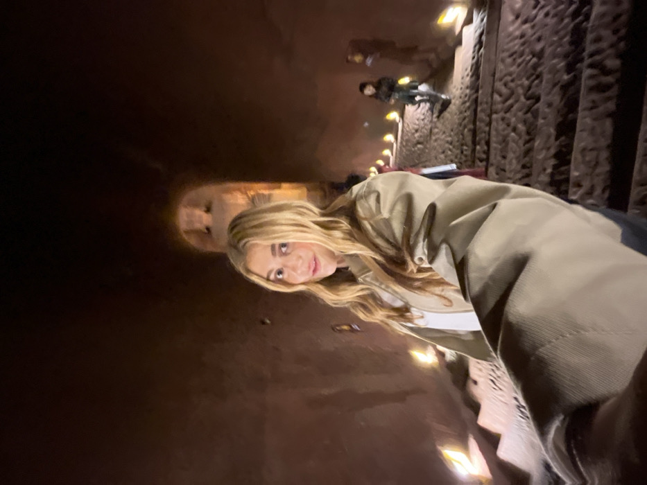 The image depicts a young adult female with long blonde hair, wearing a tan jacket over a light top, centered in a dimly lit, rough-hewn stone tunnel or cavern. She is positioned close to the camera, holding it at arm's length, and looks forward with a slight smile. The environment consists of dark, reddish-brown rock walls and a descending stone staircase with irregular, textured steps. Small, square, warm-toned lights are affixed along the walls adjacent to the steps, providing illumination. In the mid-ground, a person in dark clothing walks down the steps on the right. Further in the background, an arched opening reveals a large, carved stone structure, possibly a statue, illuminated by light emanating from beyond the archway. This scene is located within a subterranean passage of a historical or cultural site in Shizhong, China.