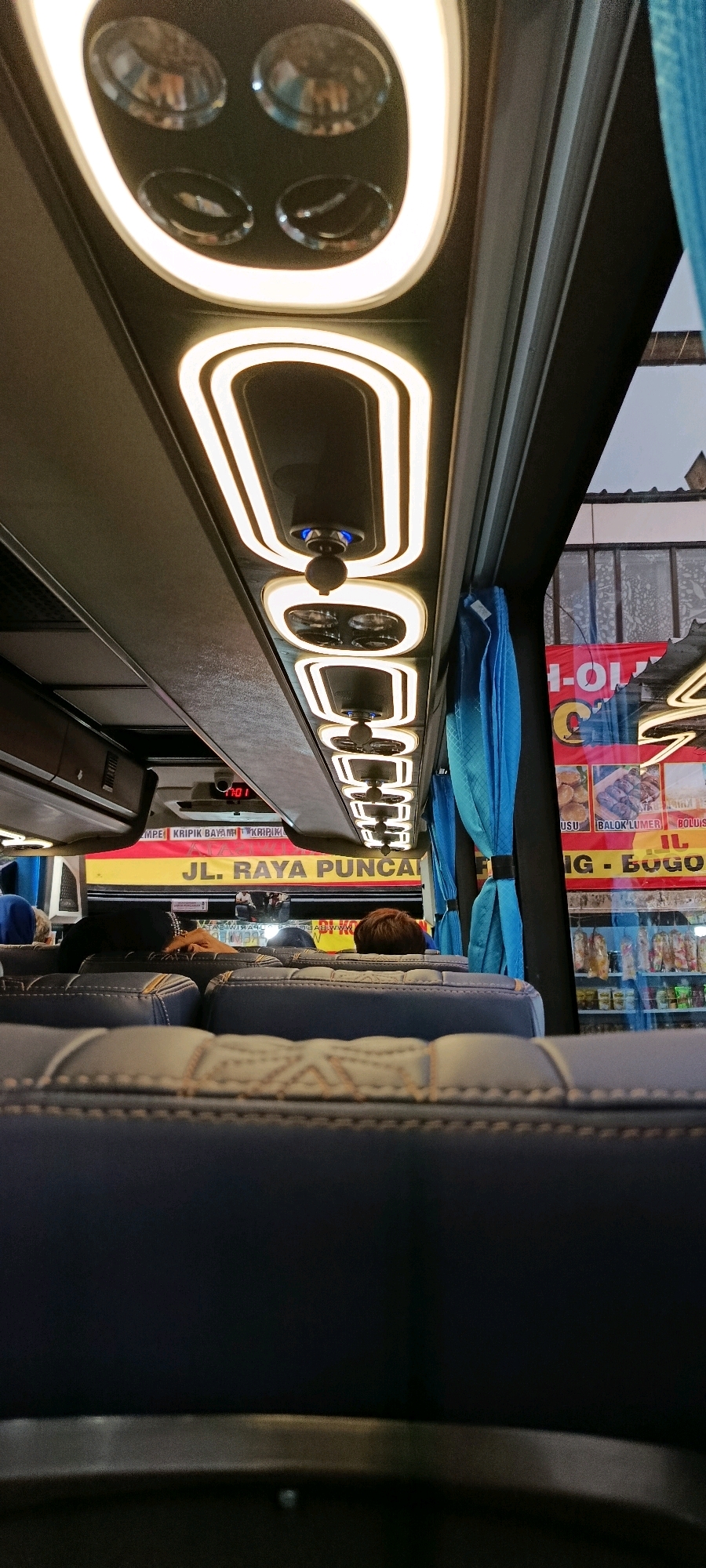 The image is an interior view of a bus, possibly in Megamendung, Indonesia, looking towards the front. The ceiling features decorative LED lighting. In the foreground, dark blue upholstered seats are visible. Further back, passengers are seated, with some appearing to be resting. Through the windows on the right, a shop or stall is visible with displays of food items and signs. One sign prominently reads "JL. RAYA PUNCAL". A digital clock on the bus ceiling displays "7:01". The overall scene suggests a journey during daytime.