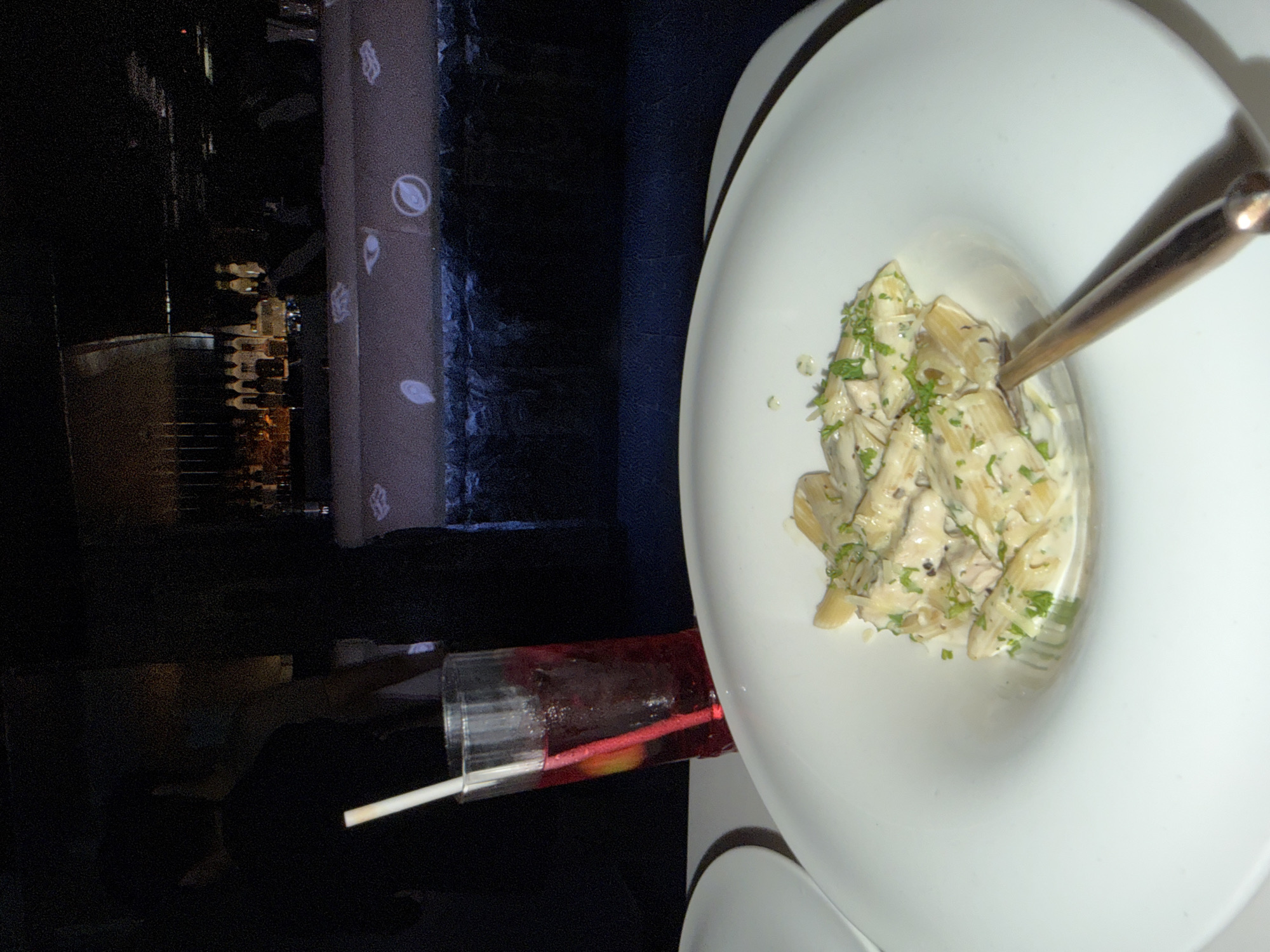 This image depicts a dimly lit indoor dining and bar area in New Delhi, India. In the foreground, a white, wide-rimmed ceramic plate contains a serving of penne pasta in a light-colored, creamy sauce, garnished with chopped green herbs. A dark metallic utensil rests within the pasta. To the left of the pasta plate, a portion of a second white plate is visible. A tall, clear glass, exhibiting condensation, holds a red beverage with a white plastic straw and a visible slice of yellow citrus fruit.
In the midground, to the left, the dark silhouette of an individual, possibly a diner, is partially visible. The background features a bar counter with a dark, textured base. The top surface of the bar counter is illuminated, displaying projected abstract symbols, including eye-like and oval shapes. Behind the bar, numerous liquor bottles are arranged on shelving, catching ambient light from the dark environment. Indistinct dark figures, consistent with staff or patrons, are present behind the bar and further in the background.