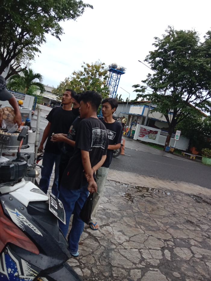 Four young men stand on cracked asphalt in front of a building with a blue roof. The building has windows, a sign, and a white door. The men are talking to each other and appear to be casually hanging out. There are trees and bushes in the background. A motorcycle with a red seat and a white license plate is parked in front of the men. The men are wearing casual clothing. The image was taken in Tugu, Indonesia, at coordinates -6.973, 110.326.