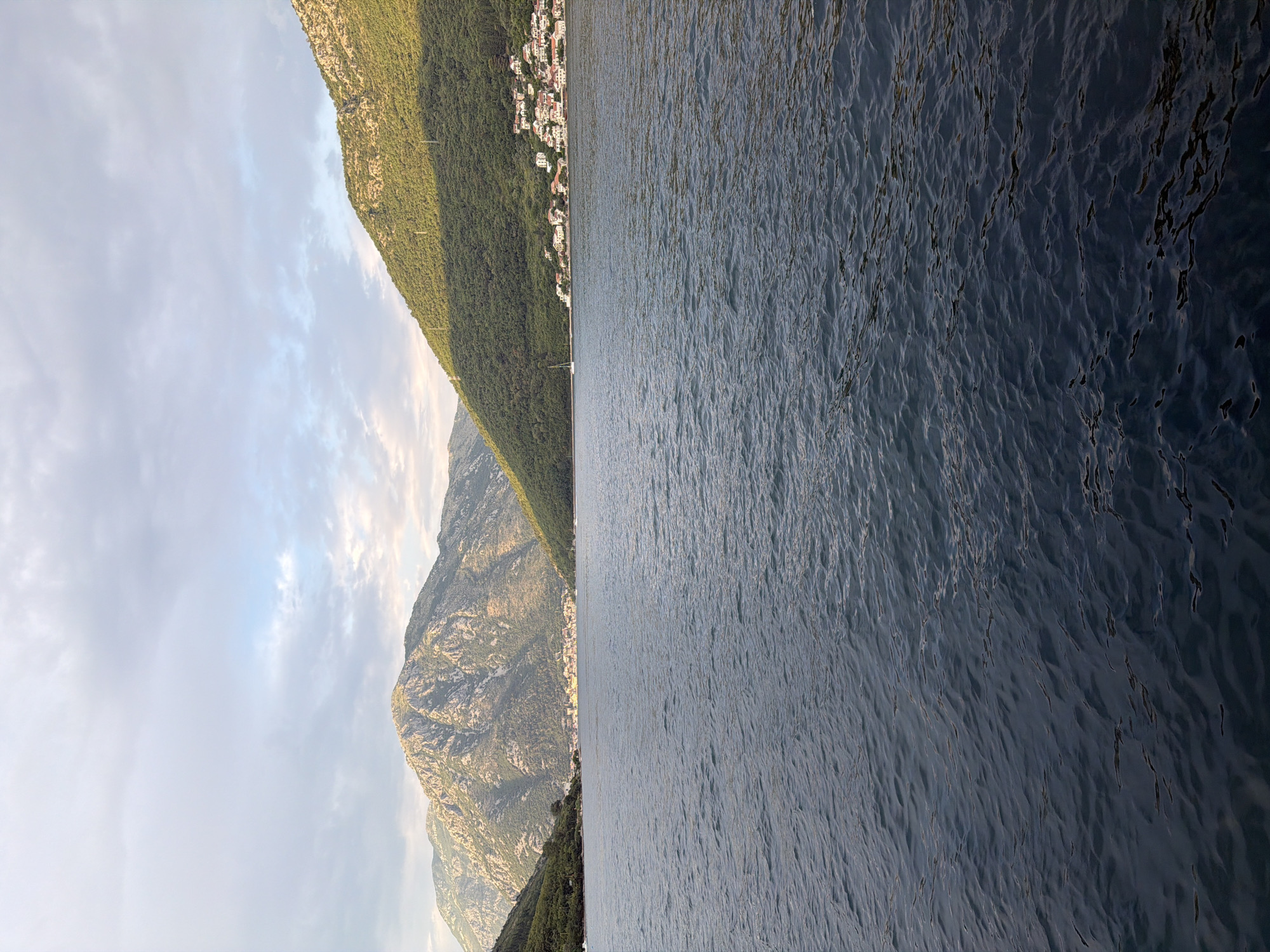 The image depicts a wide expanse of dark, rippling water in the foreground, likely a bay or fjord. In the background, verdant, tree-covered hills rise steeply on either side, meeting a hazy, overcast sky. Nestled on the slope of the right-hand hill is a cluster of white-roofed buildings, suggesting a coastal village. The architecture of the buildings, with their red-tiled roofs and white walls, is characteristic of Mediterranean settlements. A small sailboat is visible on the water near the shore. The lighting suggests either early morning or late afternoon, with shadows cast by the hills and a soft glow on the peaks. The overall impression is one of natural beauty and tranquility. While the location is specified as Đurići, Montenegro, the image itself does not contain any identifying landmarks or signs to confirm this specific location; it is a generic depiction of a coastal mountainous landscape.