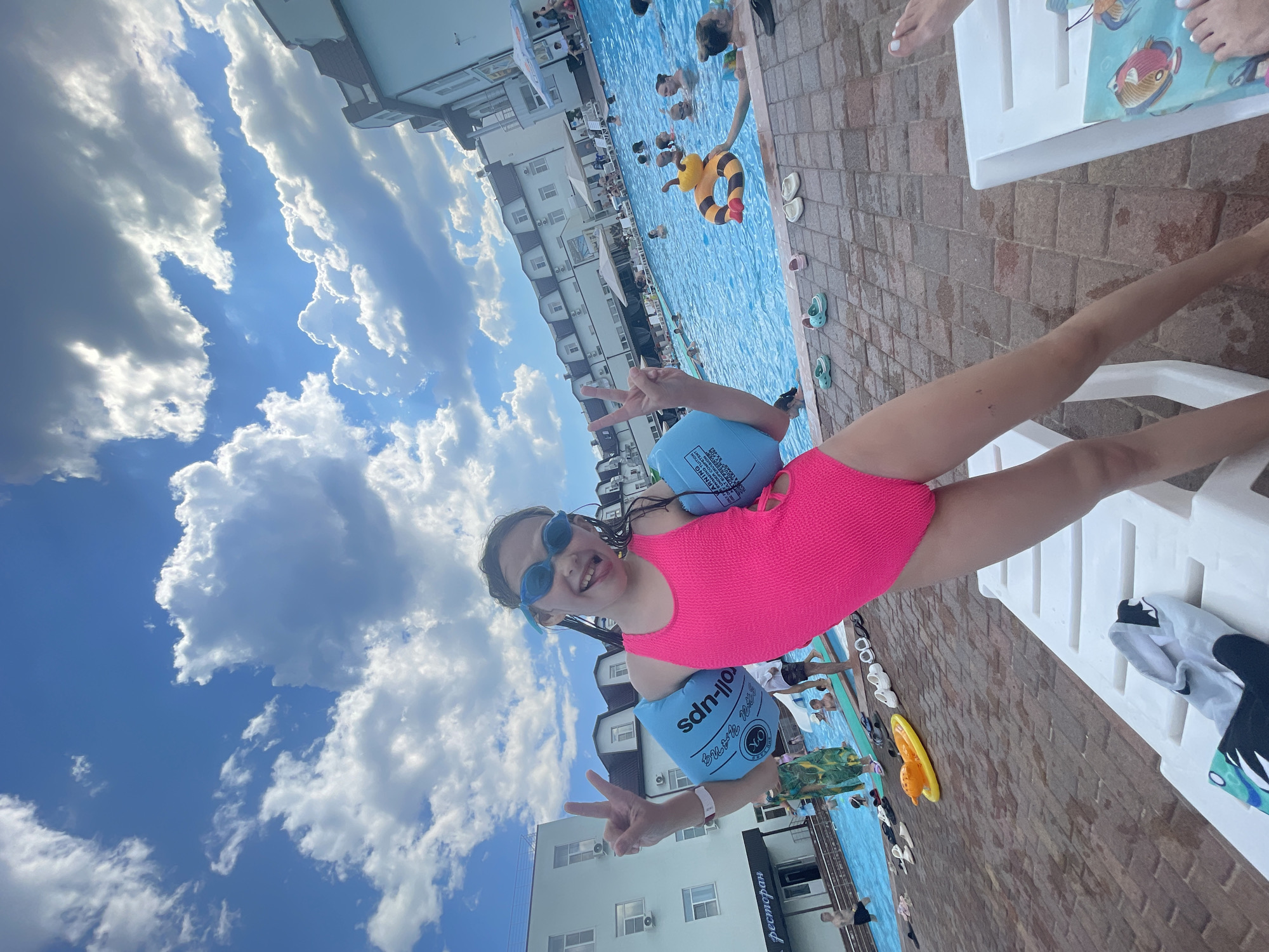 In this vertically oriented, wide-angle photograph taken at a pool in Пристанное, Russia, a young girl in a bright pink swimsuit with blue arm floaties and goggles strikes a peace sign. She is positioned in the foreground, her face turned towards the camera with a playful expression. Behind her, a crowded swimming pool reflects the bright blue sky dotted with fluffy white clouds, suggesting a sunny day.
People are visible in the pool, some swimming and others relaxing. A yellow inflatable toy shaped like a bee floats in the water. The pool area is paved with brick-like tiles and lined with white lounge chairs, one of which the girl appears to be sitting on or near. On the deck, several pairs of flip-flops are scattered, and a child's swimsuit with a shark design is draped over a chair.
In the background, a multi-story building with white walls and dark-trimmed windows can be seen, indicating a resort or hotel. The overall atmosphere is lively and suggests a summer vacation or a day spent enjoying the water. The text "ok" is visible on one of the arm floaties.