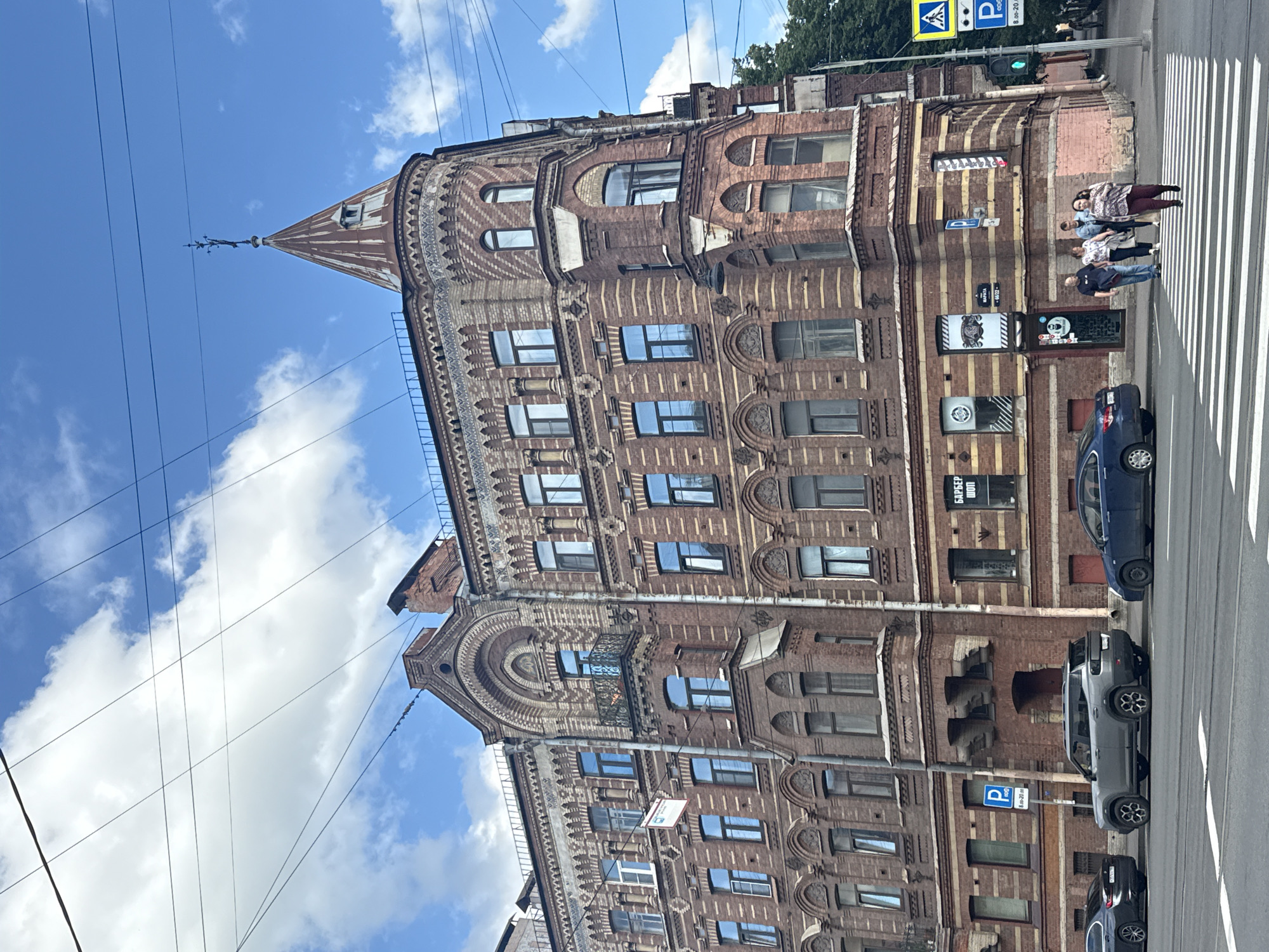 The image shows a corner building with a distinct turret in Saint Petersburg, Russia, under a partly cloudy blue sky. The building is constructed of red brick with decorative yellow brickwork and intricate architectural details, including arched windows and cornices. The turret is topped with a weather vane. In front of the building, there is a street with a pedestrian crossing and several cars parked along the roadside. Two people are walking across the pedestrian crossing, and a group of three people are walking on the sidewalk. Signs for parking and a barber shop are visible on the building's facade. Traffic lights and utility wires are also present in the scene.