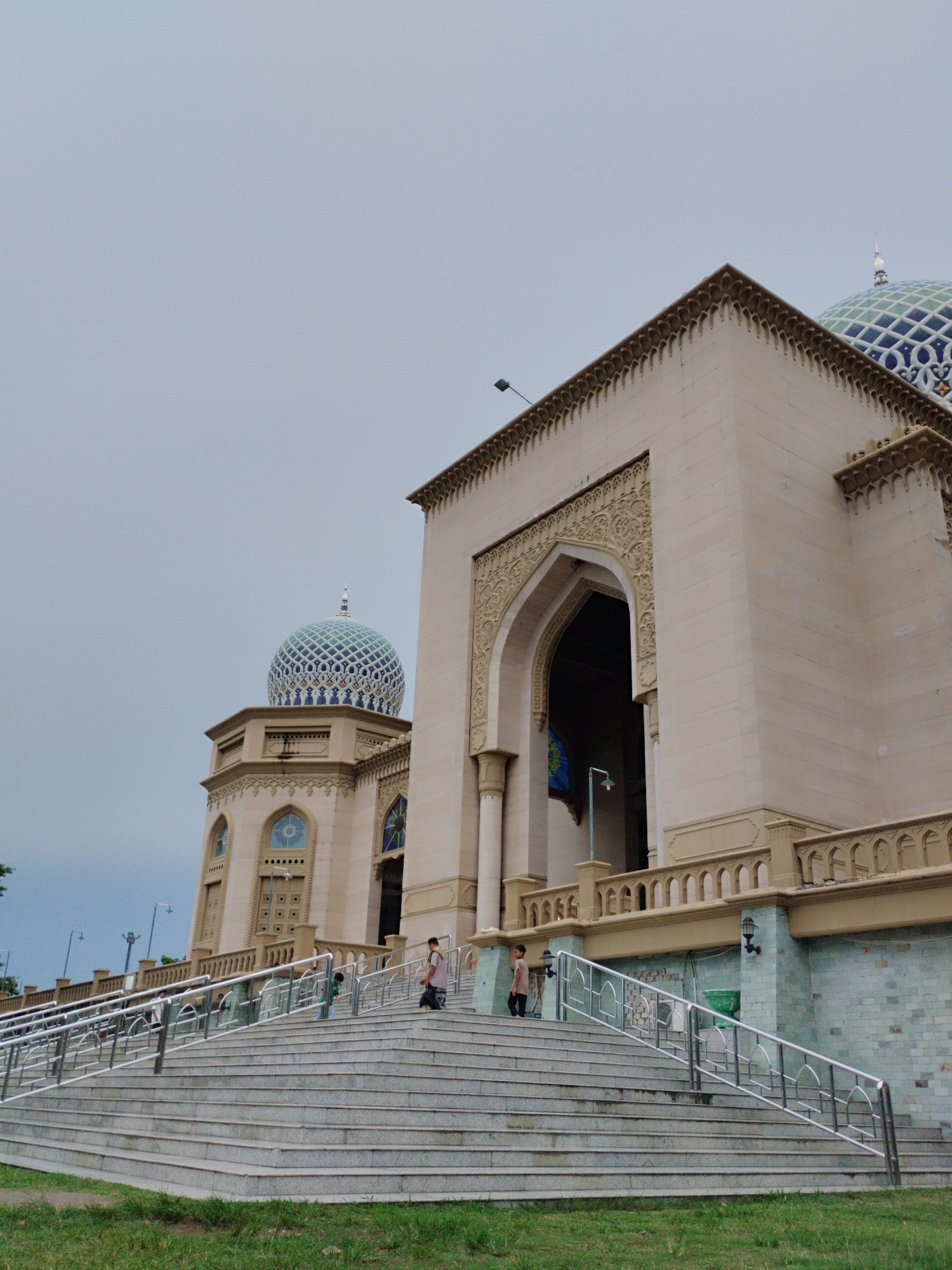The image shows the exterior of a mosque with distinctive blue and white tiled domes, located in the city of Lhokseumawe, Indonesia. A wide, multi-tiered staircase with metal railings leads up to the main entrance of the mosque. The building is characterized by light-colored stone and intricate architectural detailing, including ornate carvings around the entrance arch and decorative cornices. Several people, including children, are visible on the stairs, some appearing to be ascending. The sky is overcast, and the surrounding area includes a patch of green grass in the foreground. The architectural style suggests a modern Islamic design.