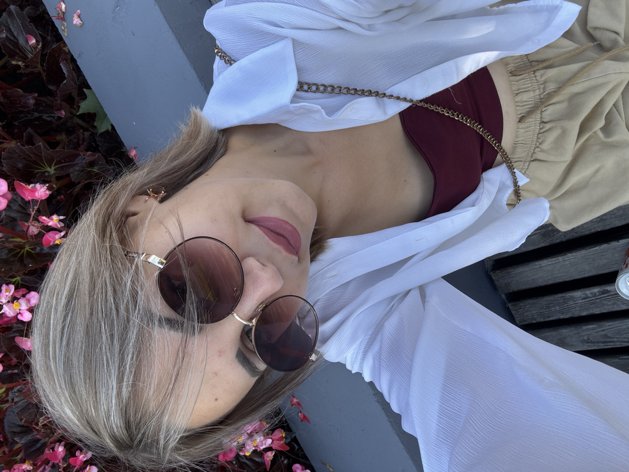 In this selfie, a young woman with blonde hair and sunglasses is partially visible, seated outdoors. She is wearing a white, unbuttoned shirt over a maroon top, and tan pants. A gold chain strap is visible across her chest. She is framed by dark leaves and pink flowers in the background. The setting appears to be a park or garden area, possibly on a bench. The Yuzhno-Sakhalinsk, Russia location is inferred from the provided context, though not explicitly visible within the image itself.