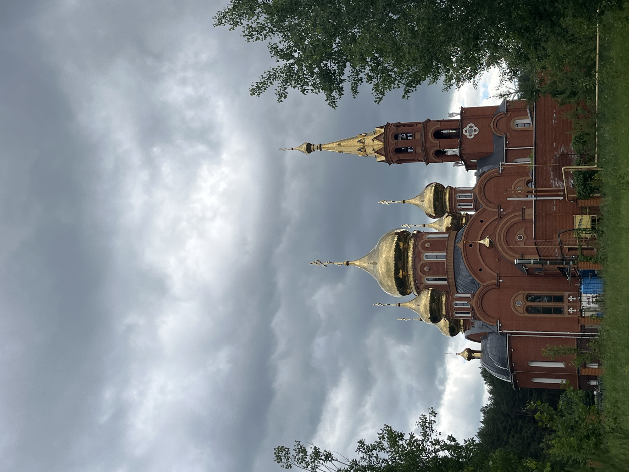 A low-angle, full shot captures a brick Orthodox church with multiple golden domes against a heavily clouded, grey sky. The church is red-brick with a central bell tower and several smaller domed structures. The foreground features lush green grass and foliage, with tall trees framing the sides of the church. A dense forest is visible in the background, behind the church. The scene appears to be set in a rural or suburban area.