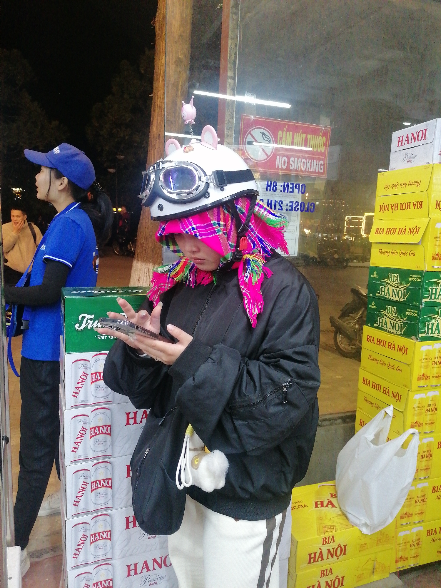 The image depicts a nighttime street scene in Sa Pa Ward, Vietnam, centered around two women standing near a storefront displaying stacks of beer boxes. In the foreground, a woman wearing a distinctive white motorcycle helmet with large goggles and a vibrant pink, green, and yellow patterned headscarf is absorbed in her smartphone. She is dressed in a black bomber jacket, white pants with a dark stripe, and carries a black shoulder bag with a white fluffy charm.
To her left, another woman in a blue uniform shirt, black pants, and a blue baseball cap stands, looking towards the left side of the frame. A man in a light-colored jacket is partially visible in the background behind the woman in blue.
The backdrop features a glass storefront reflecting neon lights and a "NO SMOKING" sign with Vietnamese text "CẤM HÚT THUỐC". Reflected numbers like "8:30" and "32:0" are also visible. On either side of the main subject, tall stacks of beer boxes are prominent. To the left, white cans and boxes are labeled "HANOI Premium BEER." To the right, yellow and green boxes are stacked, bearing the text "BIA HƠI HÀ NỘI" (Hanoi Draught Beer) and "Thương hiệu Quốc Gia" (National Brand). Some green boxes also display "HABECO" and "KIỆT TẠC". A white plastic bag rests on one of the yellow beer boxes. The artificial lighting indicates that it is dark outside.