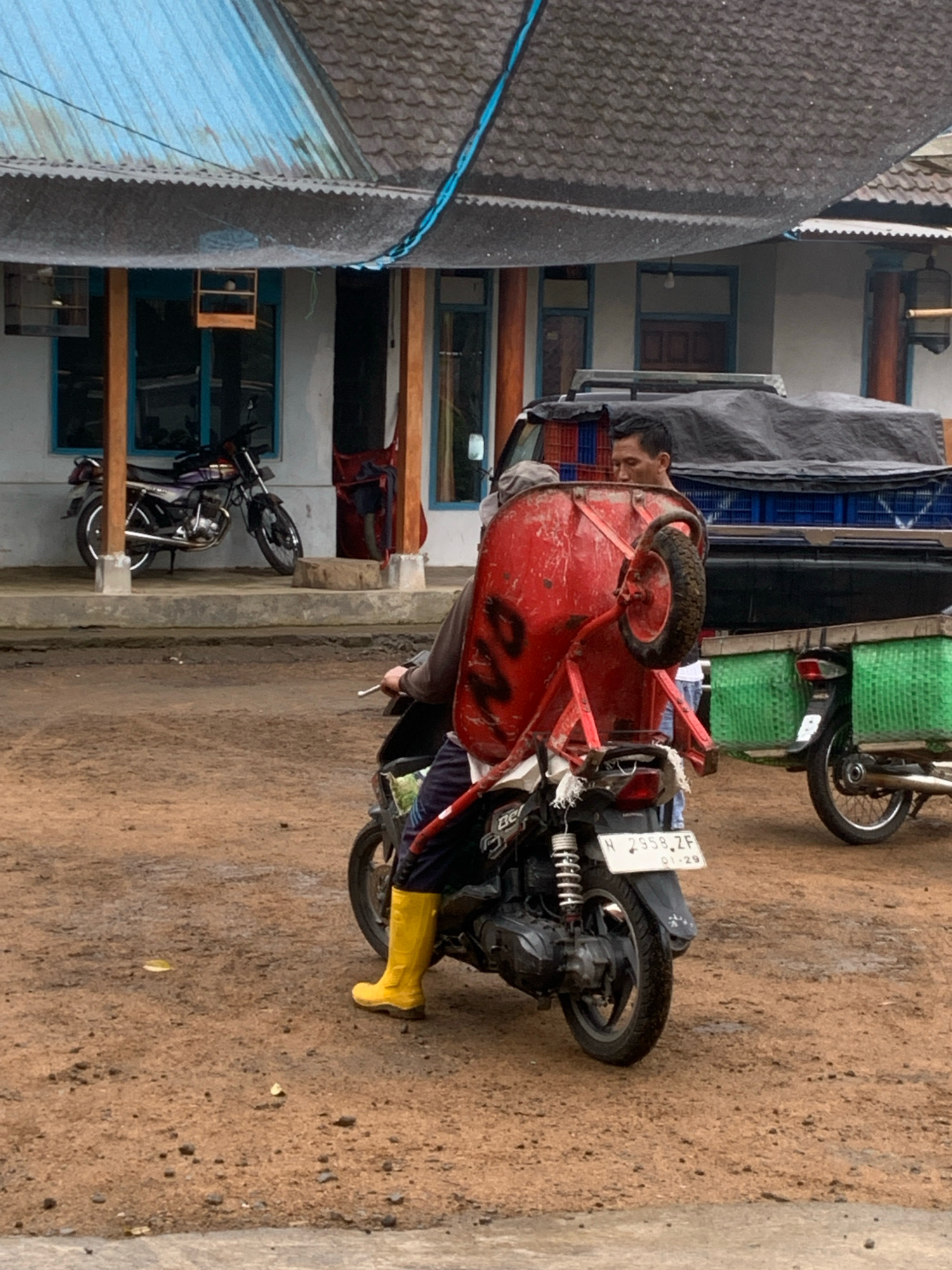 A man wearing a dark shirt and blue pants, along with bright yellow boots, is riding a motorcycle. He is transporting a large, red wheelbarrow strapped to the back of the motorcycle, with its wheel facing upwards. The wheelbarrow is secured with ropes and straps. Beside him, another motorcycle with two green crates on the back is parked. In the background, there is a building with a corrugated metal roof and blue window frames. A parked truck is also visible. The ground is dirt and appears wet in places, consistent with a rural or semi-rural environment. The license plate on the motorcycle reads "H 2958 ZF 01-29". The scene suggests a daily activity in the city of Pronojiwo, Indonesia, where such resourceful transportation methods are likely employed.
