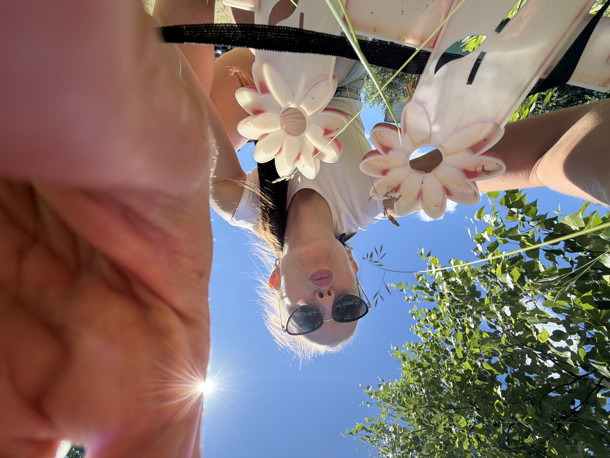 A low-angle, wide shot captures a woman outdoors on a sunny day, her face close to the camera and puckered as if to blow a kiss. She is wearing sunglasses and a white t-shirt, with her hair pulled back. She is holding up two pink and white flower-shaped plastic objects. The background is a bright blue sky, with the sun flaring in the upper right corner, and the leaves of a tree visible in the upper left. The image appears to have been taken in Birobidzhan, Russia, given the context.