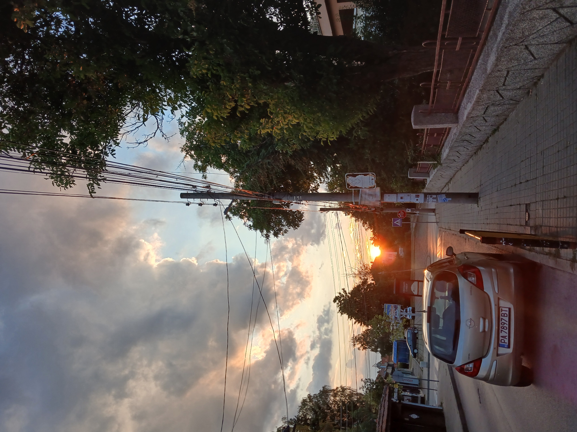This image captures a street scene in Panagyurishte, Bulgaria, during sunset. A silver Opel Corsa is parked on the right side of the road, facing away from the camera. The car has a license plate that reads "PA 7897 BT". A paved sidewalk runs alongside the street, bordered by a stone wall with a metal railing. Tall trees with green foliage line the sidewalk. Overhead, a network of power lines crisscrosses the sky. The sky itself is dramatic, with a mix of dark, heavy clouds illuminated by the warm glow of the setting sun peeking through. The sun's rays cast long shadows and create a warm, golden light across the scene. In the background, buildings and more trees are visible, some silhouetted against the bright sky. There are no people visible in the image.