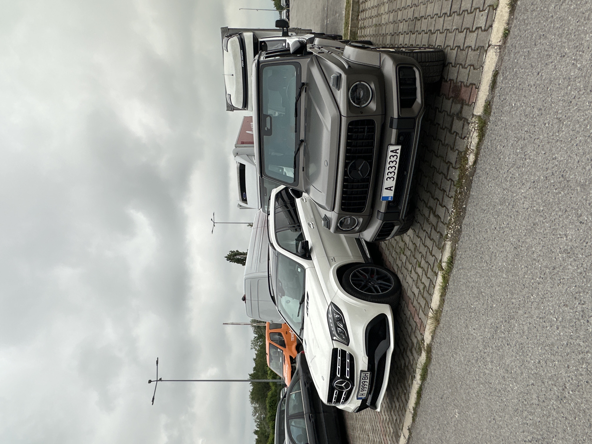 The image captures a parking area under an overcast sky, with multiple vehicles parked on a combination of interlocking pavers and asphalt. The primary subjects are two luxury Mercedes-Benz vehicles positioned prominently in the foreground.
On the left, a dark grey Mercedes-Benz G-Wagen (G-Class) is seen head-on, displaying its signature robust front grille and round headlights. Its Bulgarian license plate, "A 33333A", featuring the "BG" country code and EU flag, is clearly visible. To the right of the G-Wagen and slightly behind it, a white Mercedes-Benz SUV, likely an AMG model, is also facing forward. It features a sporty front bumper, a large central grille with the Mercedes-Benz emblem, and multi-spoke wheels. A partially visible badge on its fender indicates "V8 BITURBO". Its Bulgarian license plate reads "H 9999 BH".
In the background, several other vehicles are parked. A white semi-trailer truck is visible behind the G-Wagen, bearing the text "PONY FANTASY" on its side. Behind the white Mercedes SUV, an orange utility vehicle or van with the partial text "TEMIRAY" is parked, alongside other generic white vans and a black sedan.
The ground in the parking area is paved with grey, textured blocks, while an asphalt road runs alongside, separated by a concrete curb with some grass and weeds. The sky is uniformly light grey, filled with clouds, suggesting an overcast daytime. Several dark street light poles rise into the sky. The visible Bulgarian license plates align with the location context of Varna, Bulgaria.