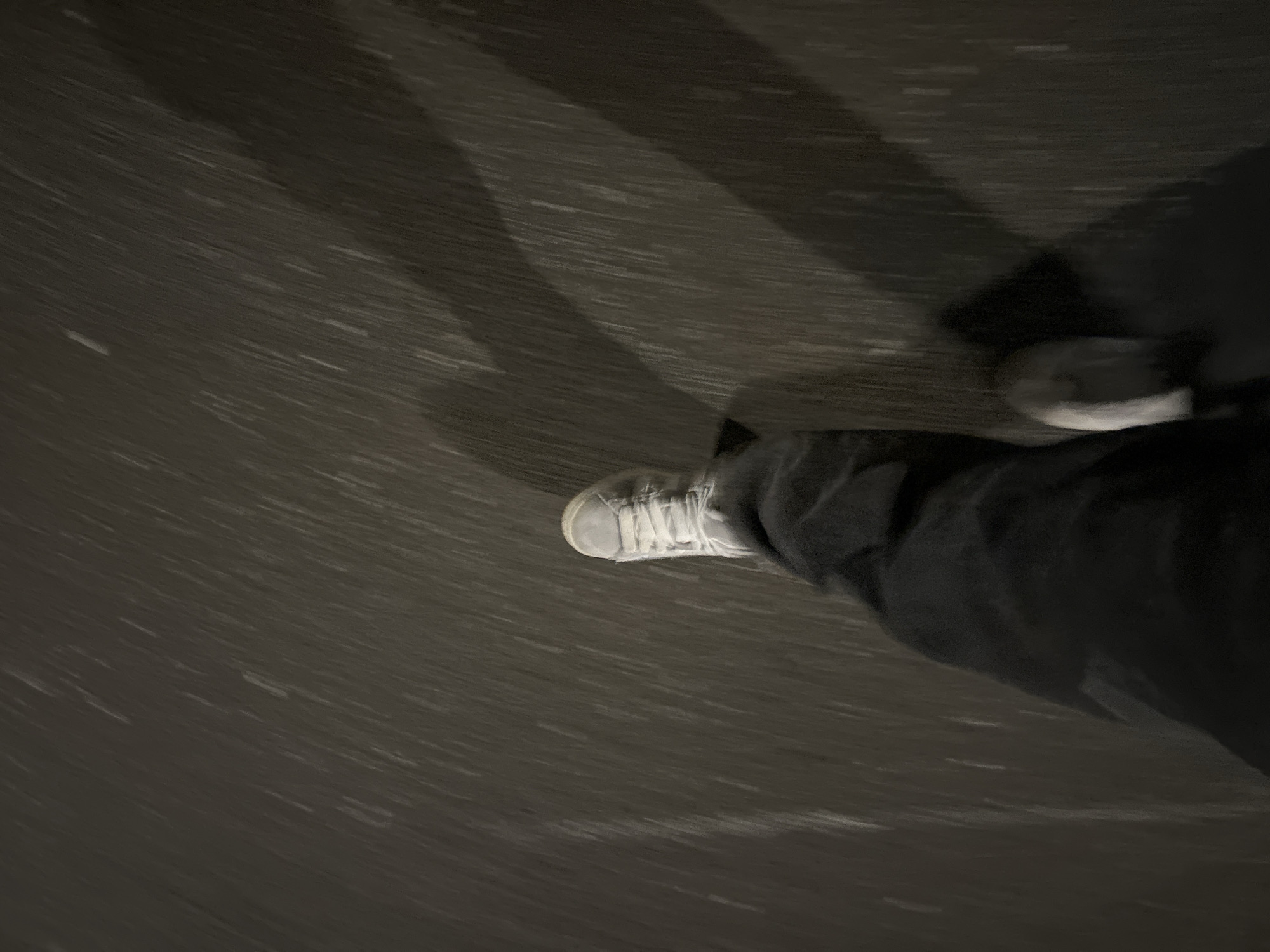 The image shows a ground-level view of a person's legs and shoes while walking on a dark surface. The surface texture suggests a road or pavement, possibly wet. The person is wearing dark pants and light-colored sneakers with white laces. The motion blur indicates movement during the photograph's capture. The location cannot be determined from this image alone.