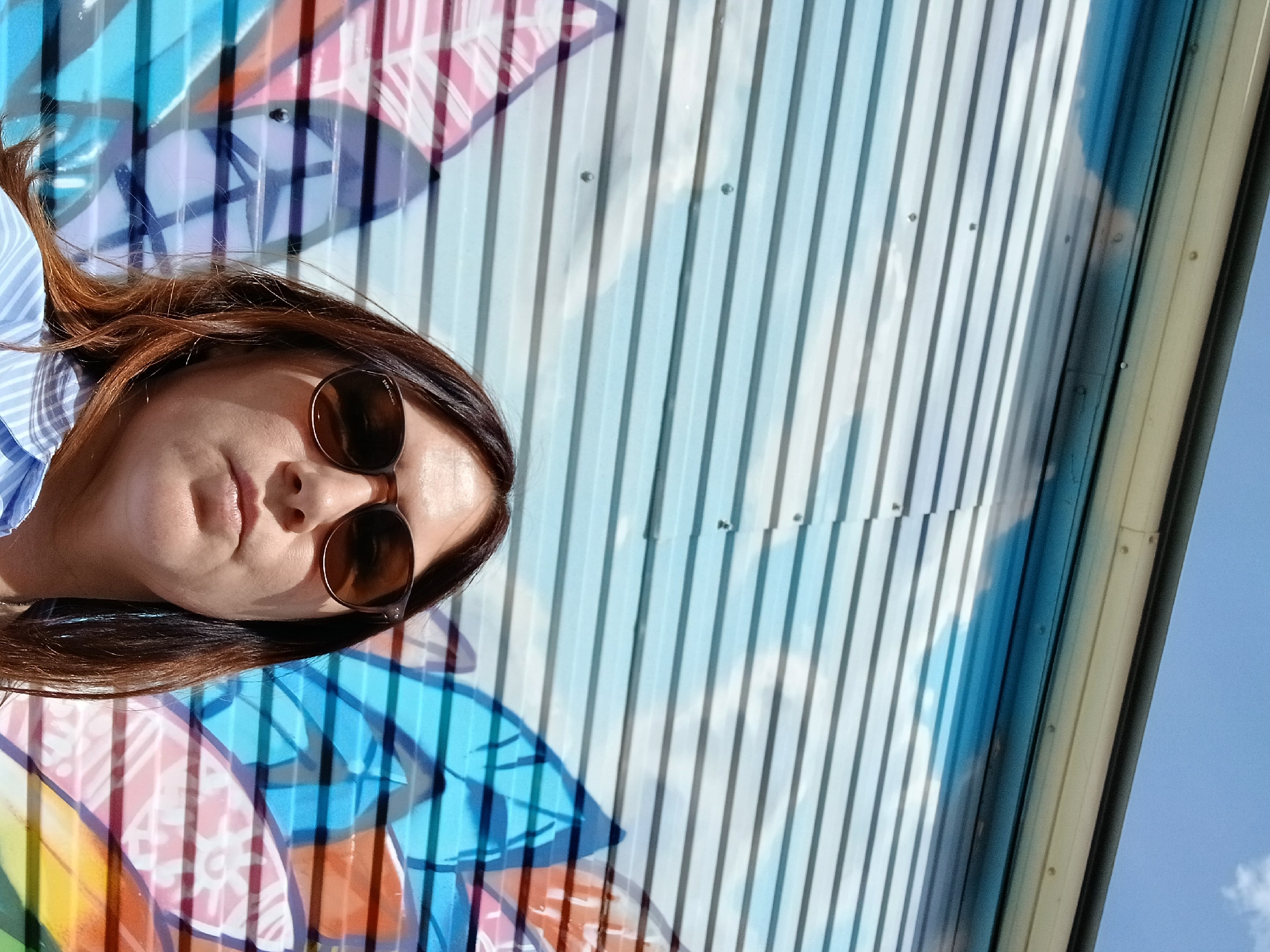 An outdoor, low-angle, medium shot shows a woman wearing sunglasses and a light blue and white striped collared shirt. She is looking directly at the camera with a neutral expression. Her long, brown hair is falling around her shoulders. Behind her is a corrugated metal wall painted with a colorful mural of abstract shapes in shades of blue, pink, orange, and yellow, and what appears to be a blue sky with white clouds. The mural is partially obscured by the horizontal lines of the corrugated metal. The top of the frame shows a glimpse of a blue sky with some white clouds. The setting is an urban environment, likely a street or alleyway.