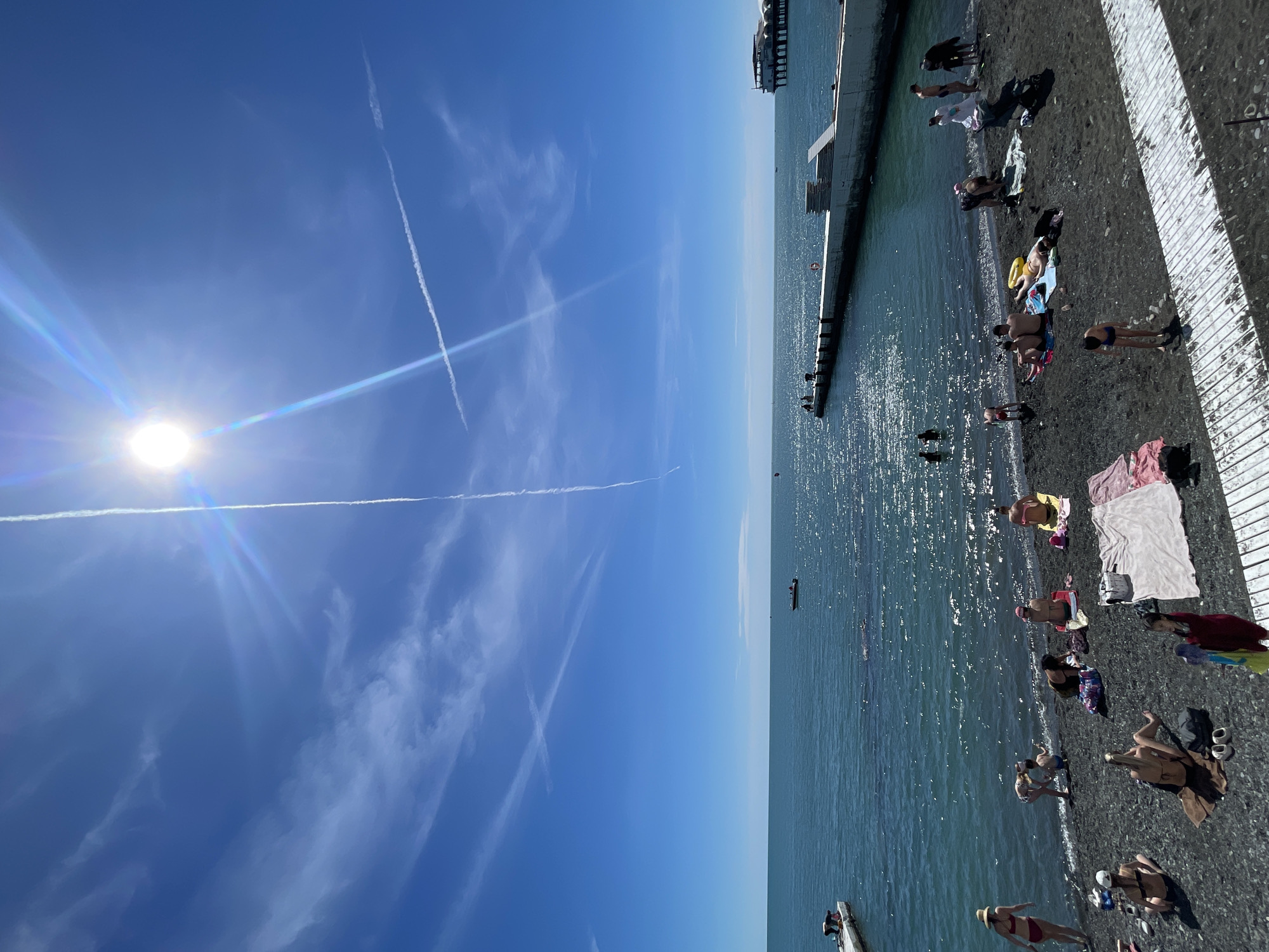 This image captures a vibrant, sunny day at a pebble beach in Sochi, Russia. The scene is split between a brilliant blue sky and the sparkling sea with a busy shoreline.
The sky, occupying the left side of the frame, is a clear azure, with the sun shining brightly in the upper left, producing a prominent lens flare and distinct sunbeams. Several white airplane contrails streak across the expanse, creating intersecting lines, and a few thin, wispy clouds are scattered about.
On the right, the deep blue-green water of the sea glistens with reflections of the sunlight, suggesting calm conditions. Concrete piers or breakwaters extend from the shore into the water, with steps leading down to the sea. A distant, multi-story building is visible along the coastline, and a small boat can be seen further out in the water.
The beach itself is composed of dark pebbles and is densely populated with people enjoying the day. Individuals are seen sunbathing on towels or directly on the beach, while others are wading, swimming, or walking along the water's edge. They are dressed in swimwear, with some wearing hats or using beach accessories like a yellow inflatable ring. The overall impression is one of leisure and relaxation under a bright mid-day sun. No discernible text is visible in the image.