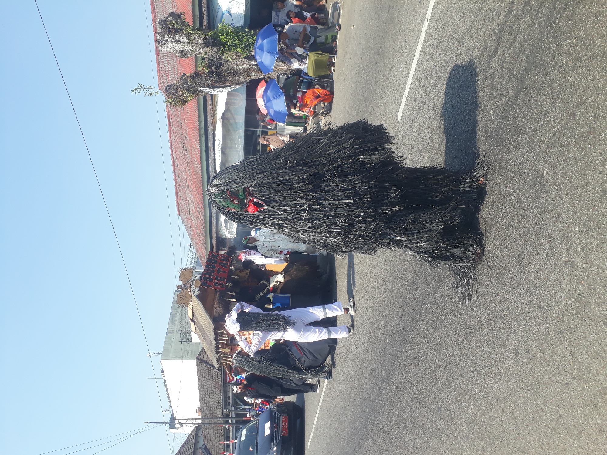 In Pangkalpinang, Indonesia, a street parade is underway under a clear blue sky. In the foreground, a person in a full-body costume resembling a shaggy, dark creature stands on the asphalt road. The costume is dense and black, with a distinct green and red face visible through the fringe. The creature's shadow is cast on the road to its right.
Further down the road, a group of people in costumes are participating in the parade. Two individuals wear long black wigs and dark robes, while another person in a white jumpsuit adjusts their white wig. Behind them, a makeshift stall or float is visible, adorned with what appear to be masks and other decorative items. A sign on the stall reads "PONDOK SETAN".
On the right side of the road, spectators are gathered, some seated on chairs and a stroller, others standing. Two blue umbrellas are open, providing shade. A black car with a license plate reading "BN 1239 TZ" is positioned on the left side of the road, with more people in the background. Buildings with red roofs line the street, and power lines are visible against the sky. The environment suggests a sunny day, likely during an event or festival.
