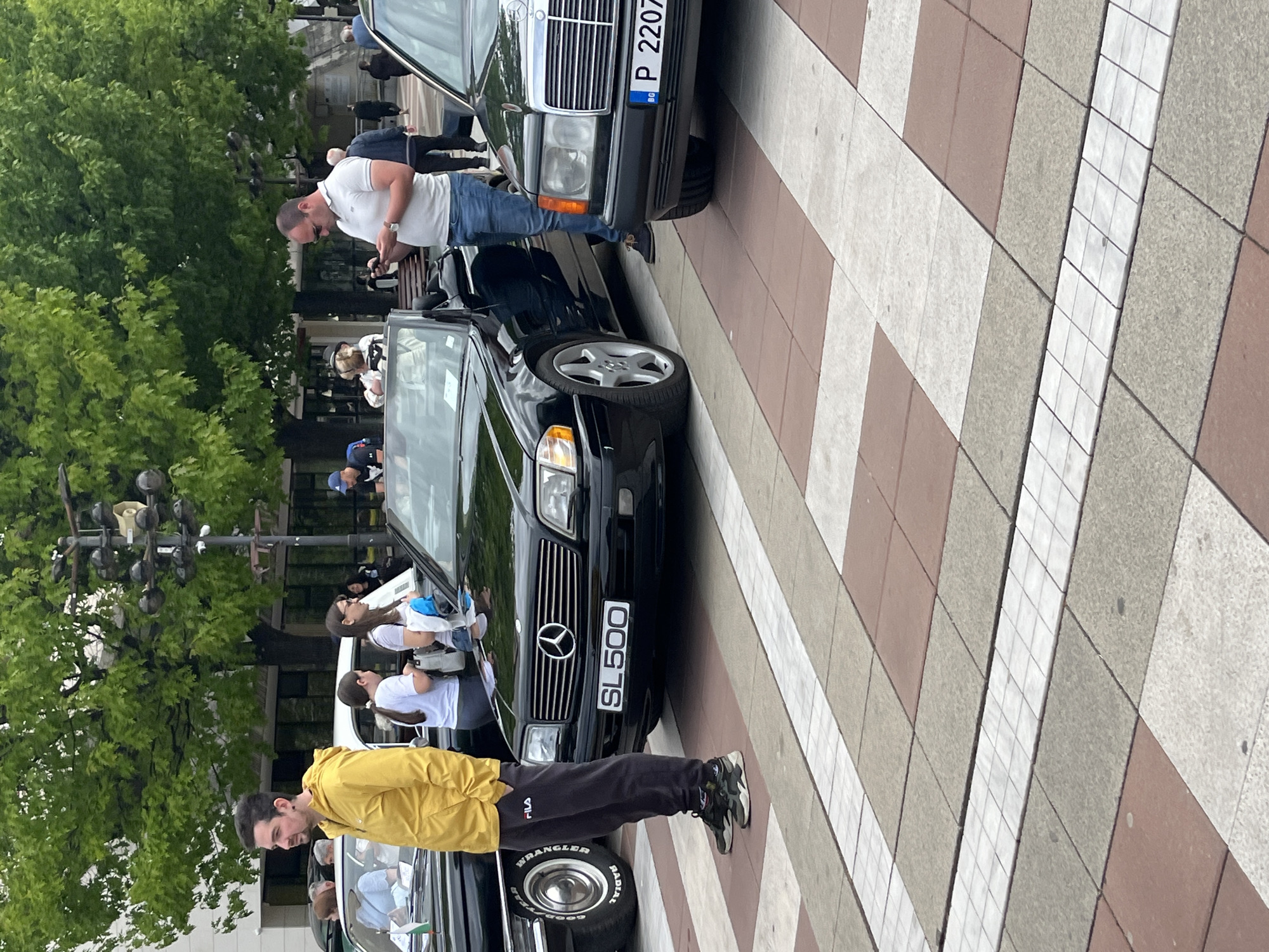 Here's a description of the image:
The image shows a street scene with several people and cars. The main focus is on a black Mercedes-Benz SL500 parked on a tiled surface. A man in a yellow jacket and dark pants is standing near the front of the SL500. Another man, wearing a white shirt and jeans, is standing near a dark-colored car with a license plate "P 2207 BG" behind the SL500. Other people are standing and walking around in the background. The environment includes trees and streetlights. The location is Unknown, Unknown.