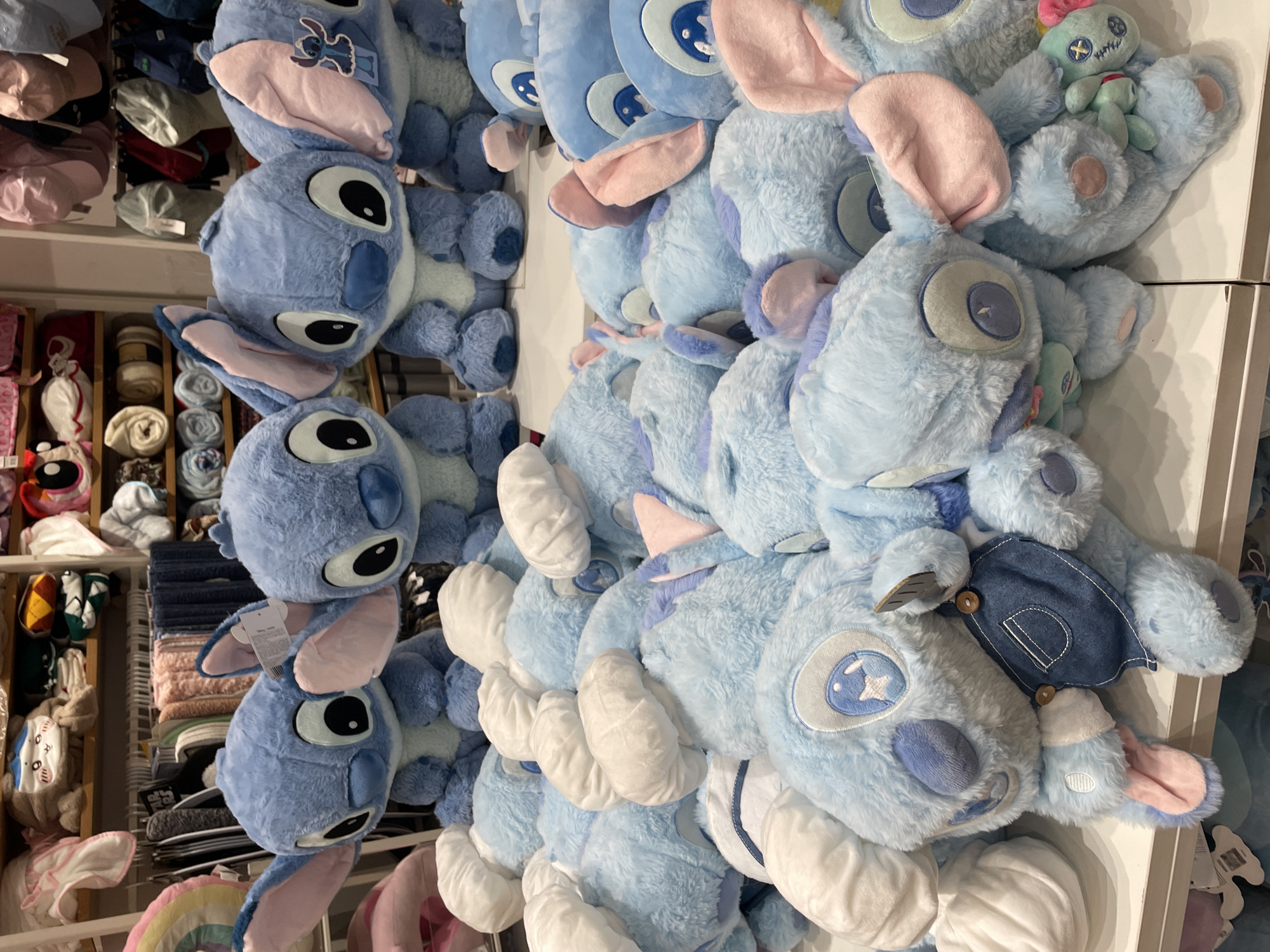 The image shows a display of numerous blue plush Stitch toys from Disney's Lilo & Stitch franchise. The toys are arranged on shelves in a retail store. In the foreground, a pile of Stitch plush toys is visible, with their characteristic large ears and blue fur. Some of the Stitch toys are wearing denim overalls. In the background, more Stitch toys are displayed on higher shelves. Behind the Stitch toys, various other retail items are visible, including folded blankets and hats on shelves. The lighting suggests an indoor retail environment. The location is identified as a retail store in Davao City, Philippines.