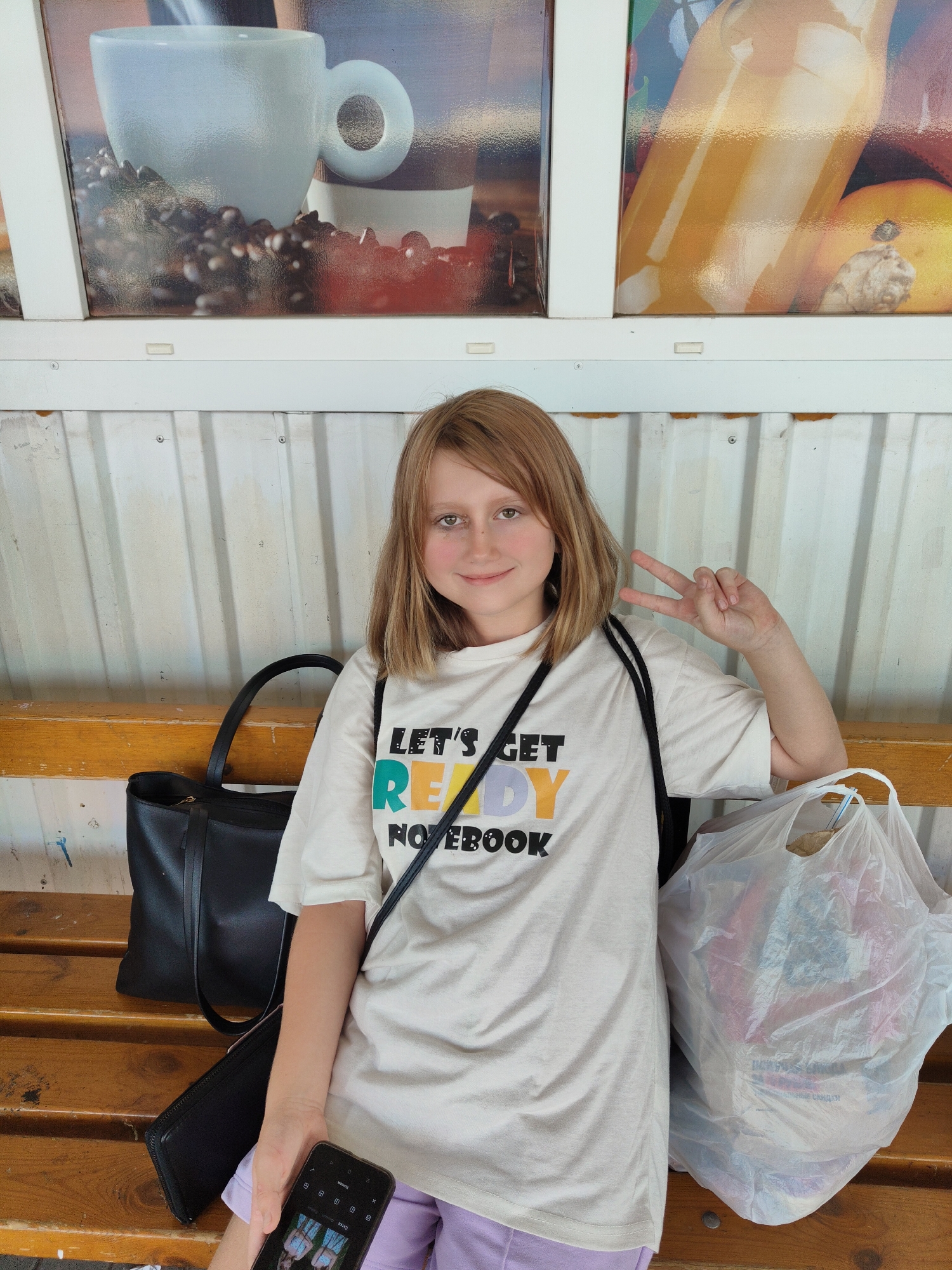 A young blonde girl with fair skin and rosy cheeks sits on a wooden bench in front of a corrugated metal wall. She wears a light-colored t-shirt with the words "LET'S GET READY NOTEBOOK" printed on it in a colorful font, and lavender shorts. She is holding a smartphone in her left hand and making a peace sign with her right hand. Beside her on the bench are a black tote bag, a black clutch, and a large white plastic bag filled with items. The background features a mural with images of a coffee cup and a glass of orange juice. The scene is outdoors, possibly in a market or cafe setting, given the mural and the bench.