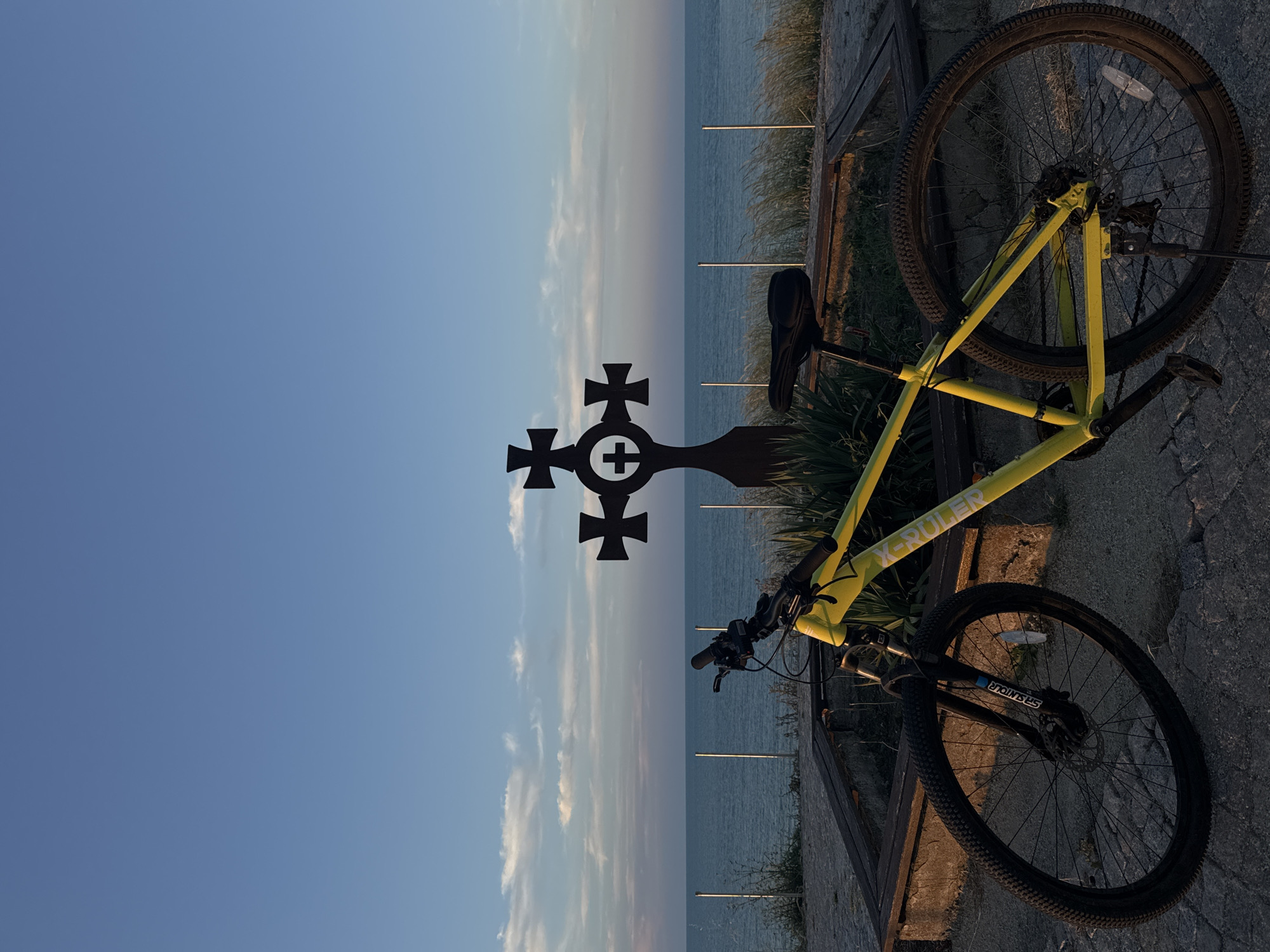 A yellow mountain bike is parked on a cobblestone surface with a wooden bench and vegetation behind it. In the background, the Black Sea is visible under a blue sky with scattered clouds. A dark, ornate cross monument stands to the right of the bike, positioned between the bike and the sea. The scene appears to be a viewpoint or memorial along the coast in Tsarevo, Bulgaria, bathed in the warm light of either sunrise or sunset. There are no people present in the image.