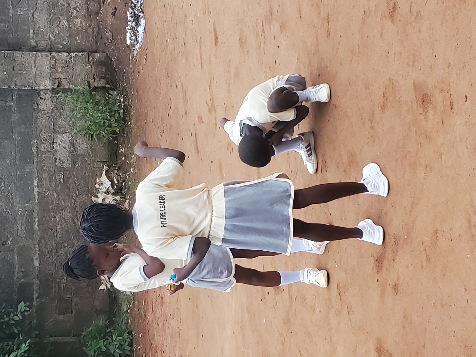 Three dark-skinned individuals are located on bare ground next to a weathered brick wall and greenery. The individuals wear light-colored shirts, with one shirt reading "FUTURE LEADER," and gray shorts with white socks and white shoes. One individual stands while another leans against them. The third individual is crouched over and appears to be facing away from the other two. The location is Ukwu-Abwa, Nigeria.