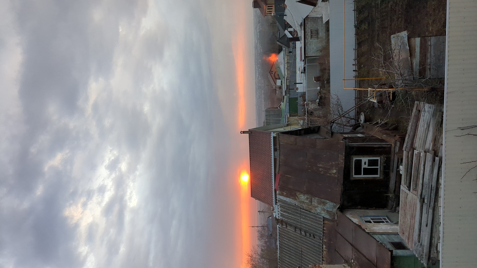 The image shows a cityscape at sunset in Ulyanovsk, Russia. The sky is filled with gray clouds and the sun is visible on the horizon, casting an orange glow over the landscape. Buildings are scattered throughout the scene, including residential houses with rusty roofs and windows. In the foreground, there is a dilapidated building with stacks of wooden planks leaning against its side. The environment appears to be a mix of urban and rural, with trees and vegetation interspersed among the buildings.