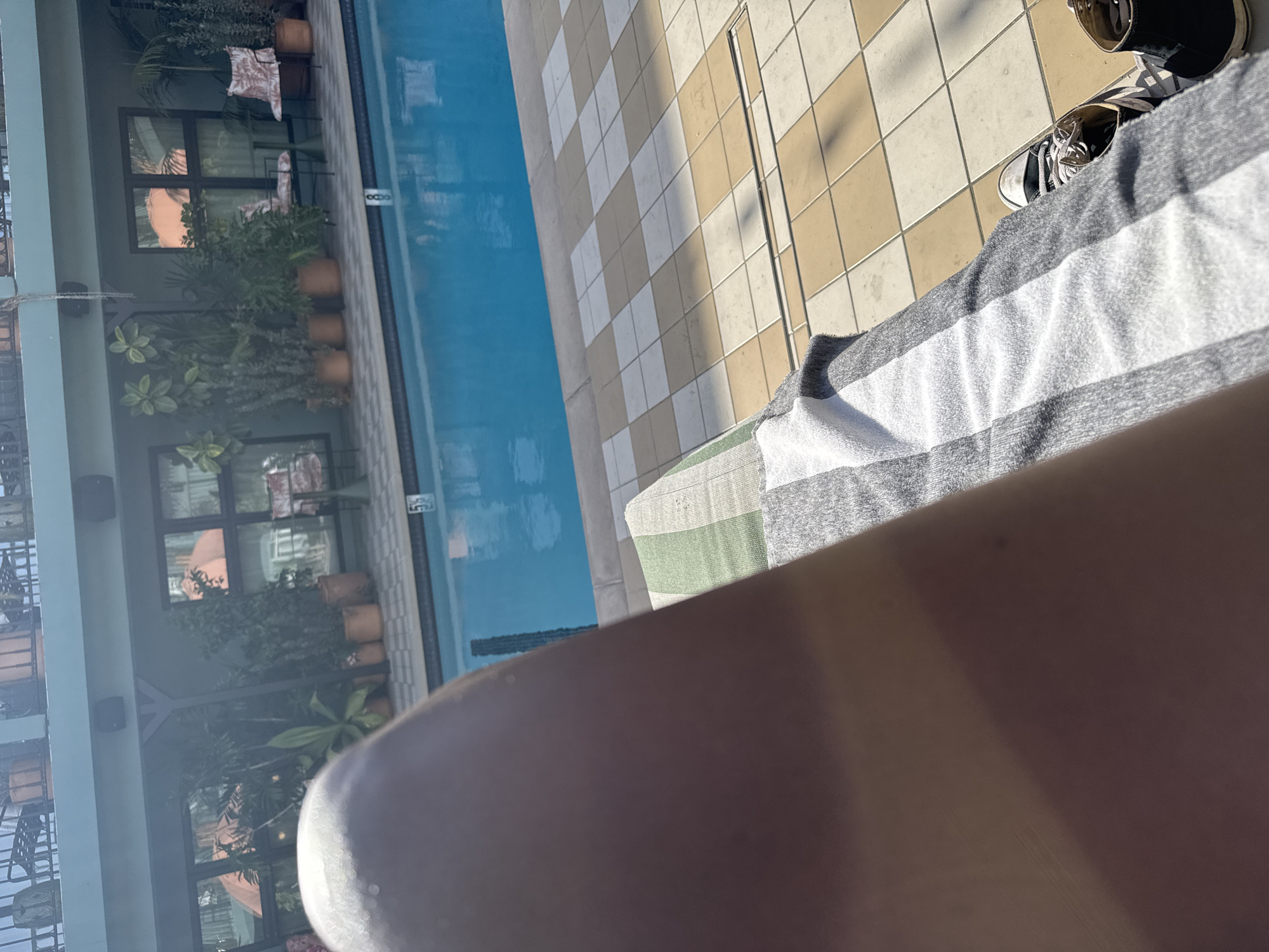 The image captures a first-person perspective of a relaxing scene by a swimming pool in West Hollywood, United States of America, under bright, sunny conditions. The lower right portion of the frame is dominated by a person's sunlit arm or leg, partially obscured and blurred. Next to it, a striped towel with grey and white bands is visible, with a smaller section of green and white stripes underneath. A pair of black and white canvas shoes, resembling Converse sneakers, are placed on the tiled ground to the right, suggesting the person is lounging.
The foreground features a clear blue swimming pool running along the left side, with subtle reflections on its surface. The adjacent pool deck is paved with a checkered pattern of alternating light brown and light grey or white square tiles, upon which long shadows are cast, indicating bright daytime sun.
In the background, a modern building with multiple large, dark-framed windows is visible. These windows reflect parts of the outdoor environment and possibly interior spaces, showing hints of furniture like chairs and cushions. Numerous green potted plants are arranged along the base of the building and on various ledges, enhancing the serene atmosphere. Black speakers are mounted on the exterior walls of the building. A small white marking, possibly "55" or "SS," can be seen on the inner wall of the pool near the edge. The overall scene conveys a peaceful and leisurely setting.