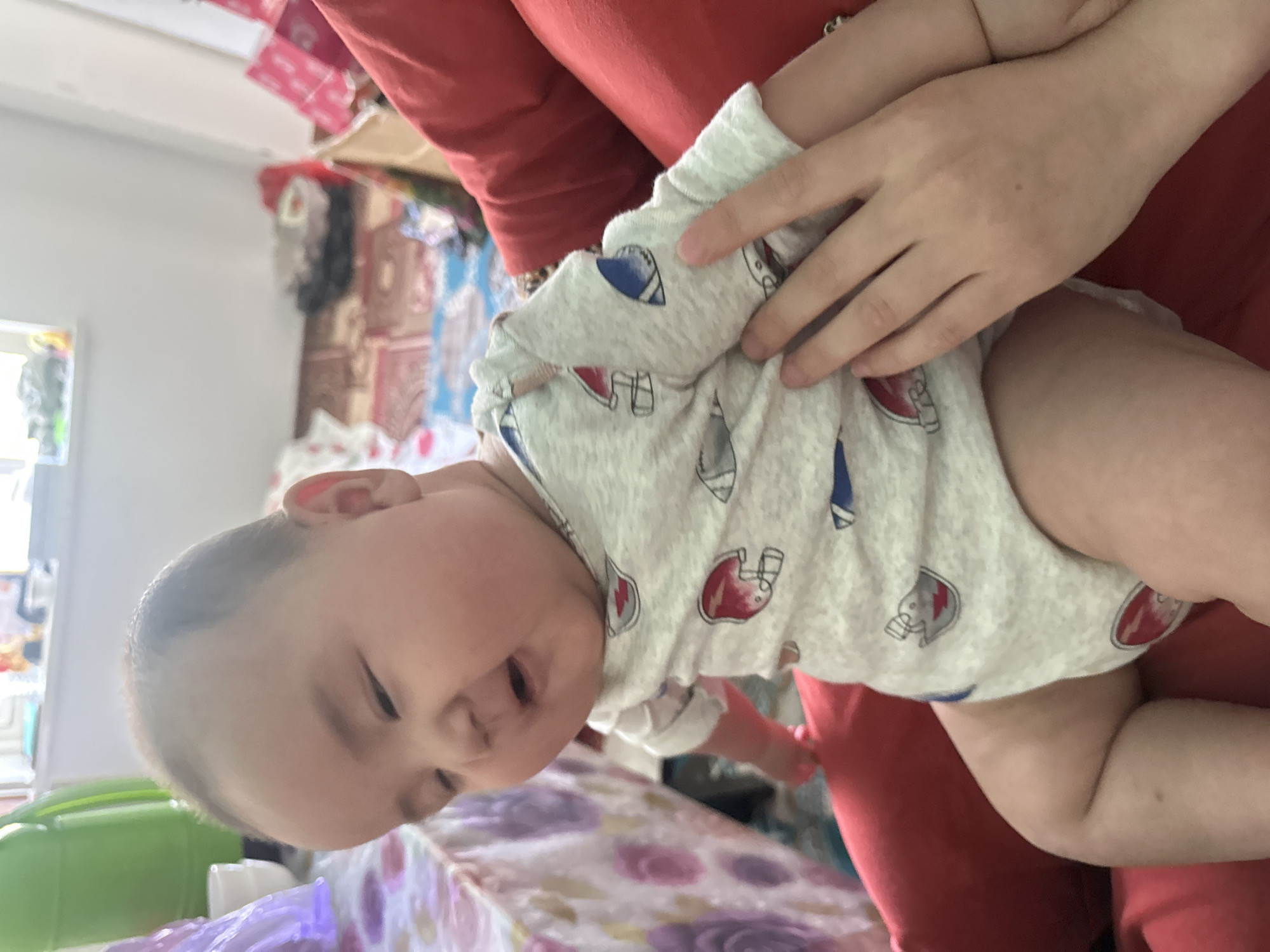 A baby wearing a light-colored onesie with a pattern of football helmets is being held by a person wearing a red shirt. The baby's head is tilted, and their eyes are partially closed. The background is blurry, but it appears to be an indoor setting with some items stacked on shelves. The lighting suggests it is daytime. The overall mood is calm and intimate, suggesting a moment of quiet bonding between the baby and the caregiver.