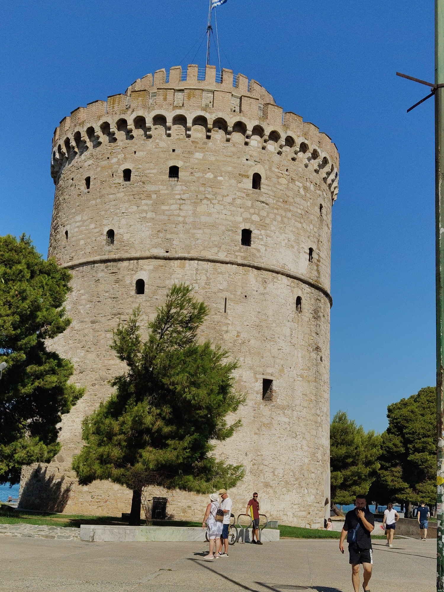 The White Tower, a prominent landmark in the city of Thessaloniki, Greece, stands tall against a clear blue sky. The ancient stone structure, circular in shape, is adorned with crenellations and small, dark windows. A Greek flag flies from its summit. In the foreground, a paved walkway stretches out, with several people walking. To the left, lush green trees partially obscure the base of the tower, and the sea can be glimpsed beyond. The overall scene depicts a sunny day in a historic urban setting.