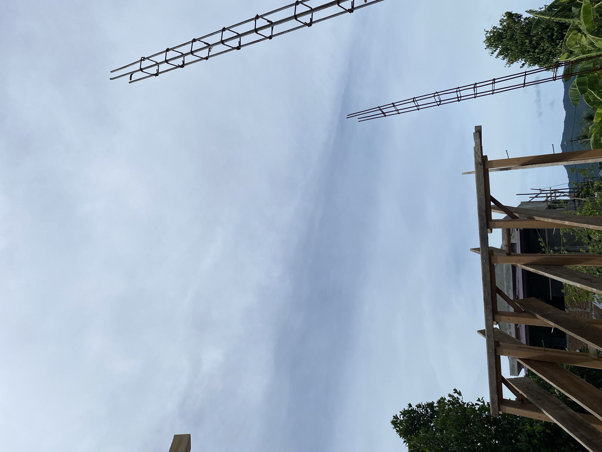 This is a photograph taken outdoors in Banda Aceh, Indonesia, capturing a construction site under a cloudy sky. The scene is dominated by the skeletal framework of a building under construction, with exposed wooden beams forming angled supports and a horizontal beam at the top. Two vertical rebar structures, resembling unfinished ladders, rise into the sky on the right side of the frame. In the background, lush green trees and banana leaves are visible, along with distant hills. The overall impression is one of ongoing development in a tropical environment.