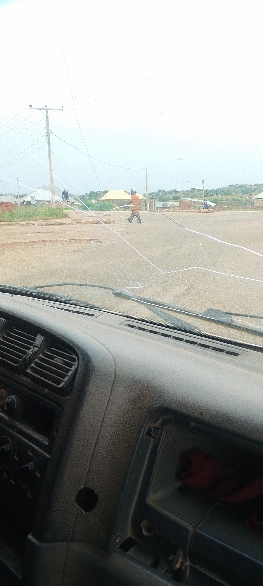 This image captures a scene from inside a vehicle in what appears to be the city of DU, Nigeria. The foreground is dominated by the interior of the vehicle, showing a portion of the dashboard with air vents and a dark textured material. Through the windshield, a man in an orange outfit is seen walking across a dusty, unpaved road. The road is marked with white tape in several places, suggesting some form of activity or construction is taking place. In the background, there are several small structures, some with corrugated metal roofs, and a utility pole with multiple wires. The sky is overcast, and the overall impression is of a rural or developing area. The image is taken from a low angle, as if from the driver's perspective.