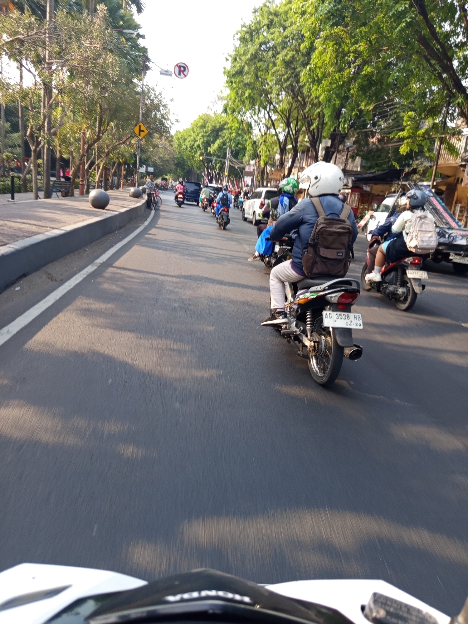 The image captures a street scene in the city of Kediri, Indonesia, likely during daylight hours given the strong shadows. The perspective is from the viewpoint of a rider on a motorcycle, looking forward along a busy road. Several other motorcycles are visible, forming a line of traffic. One motorcycle in the foreground, with a license plate reading "AG 3538 NB", carries a rider wearing a blue jacket and a brown backpack. Behind this motorcycle, another rider on a motorcycle is visible, also wearing a backpack. On the left side of the road, there is a sidewalk with trees, benches, and spherical bollards. A traffic sign with a red circle and a line through the letter "R" indicates a no-parking zone. The road itself is asphalt, with a white line marking the edge of the lane. In the background, more trees and buildings line the street, and other vehicles, including a white car and a truck, are present. The overall impression is of a typical urban traffic environment in Indonesia.
