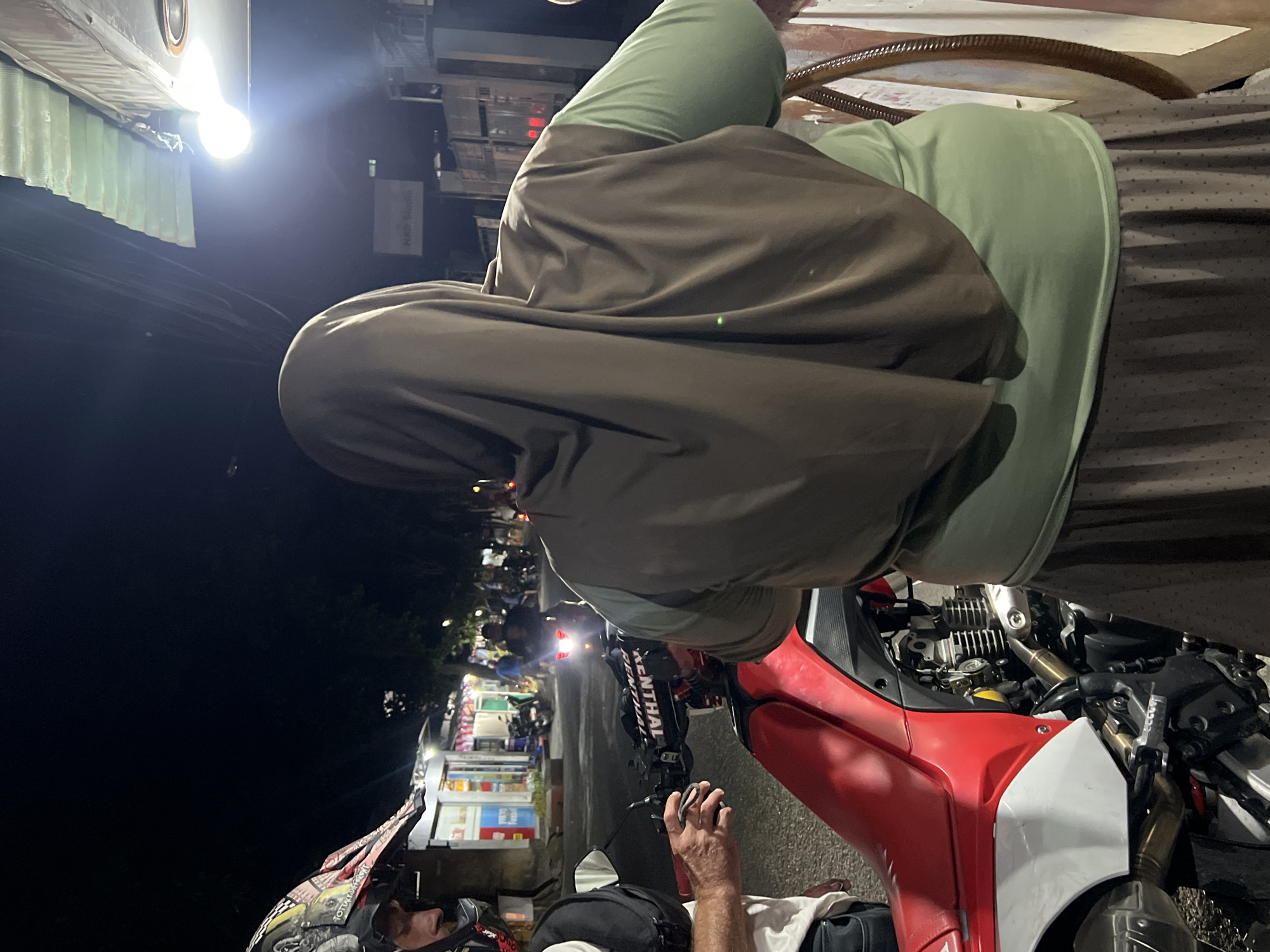 The image captures a scene at night on a street in Denpasar, Indonesia, likely near a food stall or shop, indicated by the illuminated storefronts in the background. A red and white motorcycle is partially visible in the foreground, with its engine exposed. A person wearing a motocross helmet is standing to the left, looking down at the motorcycle. To the right, a person wearing a brown hijab and a green shirt is standing, with their back to the camera. The person's outfit suggests they might be female. The street appears to be paved, and other motorcycles and individuals can be seen in the middle ground, suggesting a public area with some activity. The lighting is dim, characteristic of nighttime, with bright artificial lights illuminating some areas of the street and storefronts.