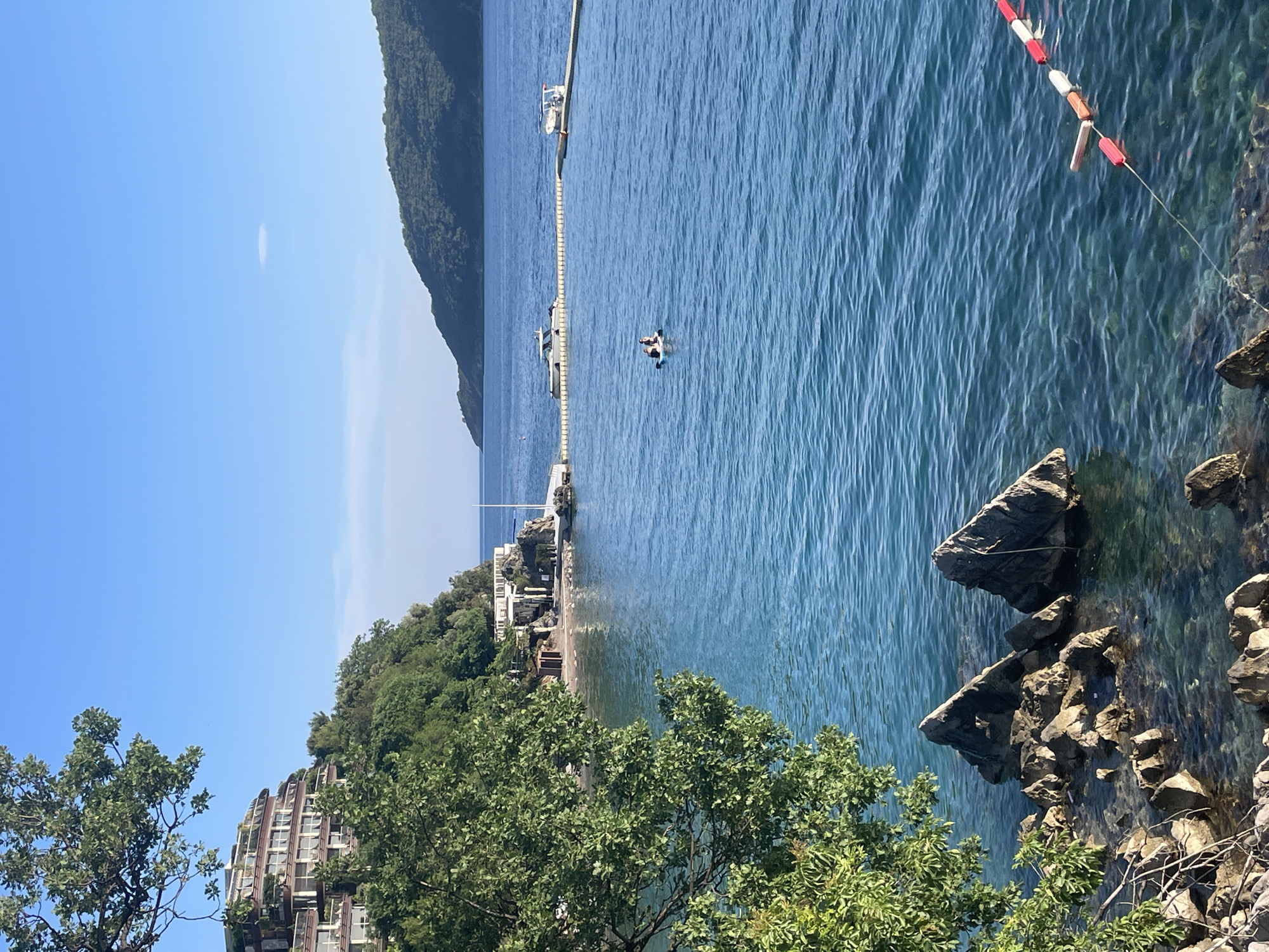 The image shows a coastal scene in Budva, Montenegro on a clear day. The blue sea occupies the majority of the frame, with rocky outcroppings in the foreground. A line of red and white buoys runs parallel to the rocky shoreline. In the water, a person is on a paddleboard next to a yellow barrier that spans the water and a docked motor boat. Beyond the water are a strip of land covered with green trees, buildings, and other structures, with a beach. A wooded peak rises on the left side of the frame. The sky is a clear blue with a sliver of cloud.
