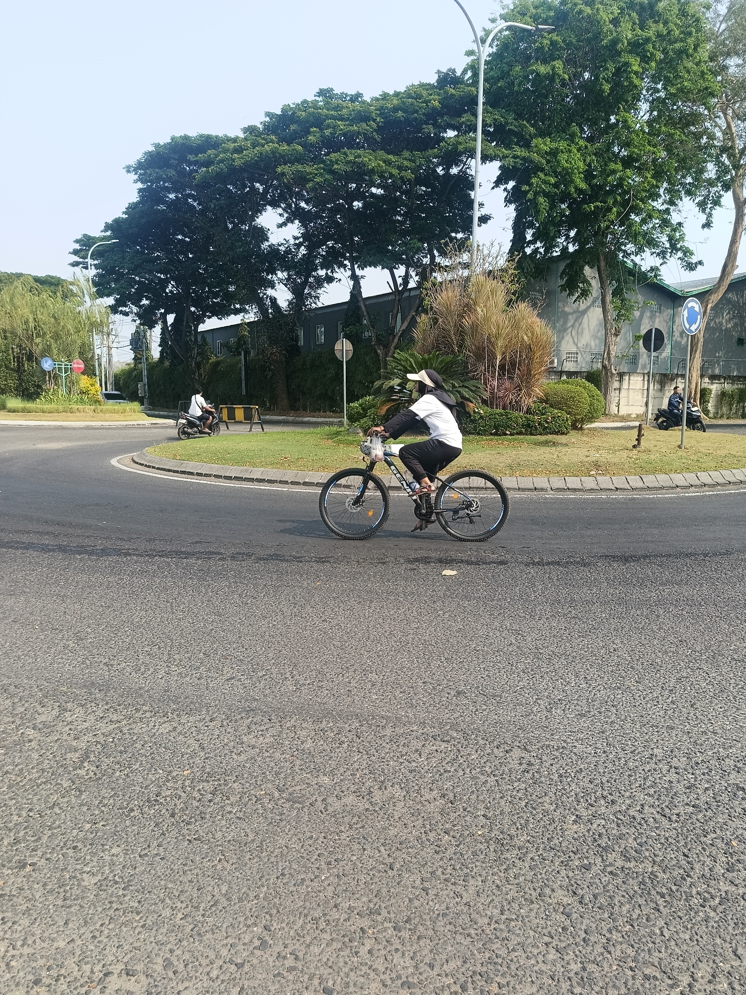 A person is riding a bicycle on an asphalt road. The person is wearing dark pants, a white shirt, a dark headscarf, and a white visor. They are holding a clear plastic bag in their left hand. The bicycle is dark colored with visible spokes. The road curves to the left. In the background, there is a grassy median with a few small trees and bushes. Further back, there is a gray building with windows, and a street light pole. There are also other vehicles and trees visible in the distance. The sky is clear and light blue. This scene appears to be in a developed area, possibly an industrial or commercial zone, given the building and manicured landscaping. The location is Calukan RT 1 RW 5 Keboansikep, Indonesia.
