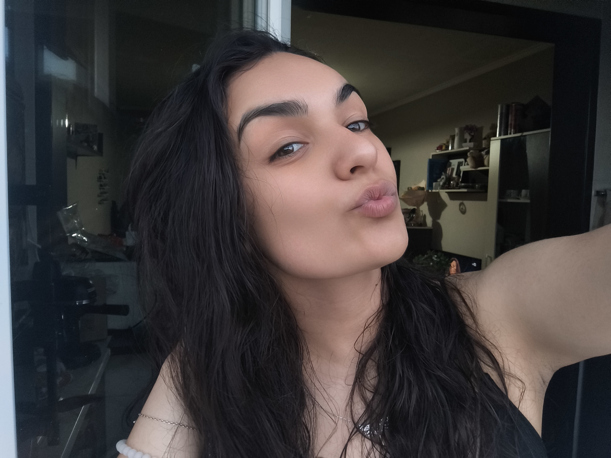 An outdoor, close-up, selfie-style photograph shows a young woman with dark hair and light skin. She is pouting, with her lips pursed forward. Her eyes are open and looking at the camera. She is wearing a black top. The background is blurred, but appears to be a room with shelves of items. The overall lighting suggests it is daytime. The image is located in Sofia, Bulgaria.