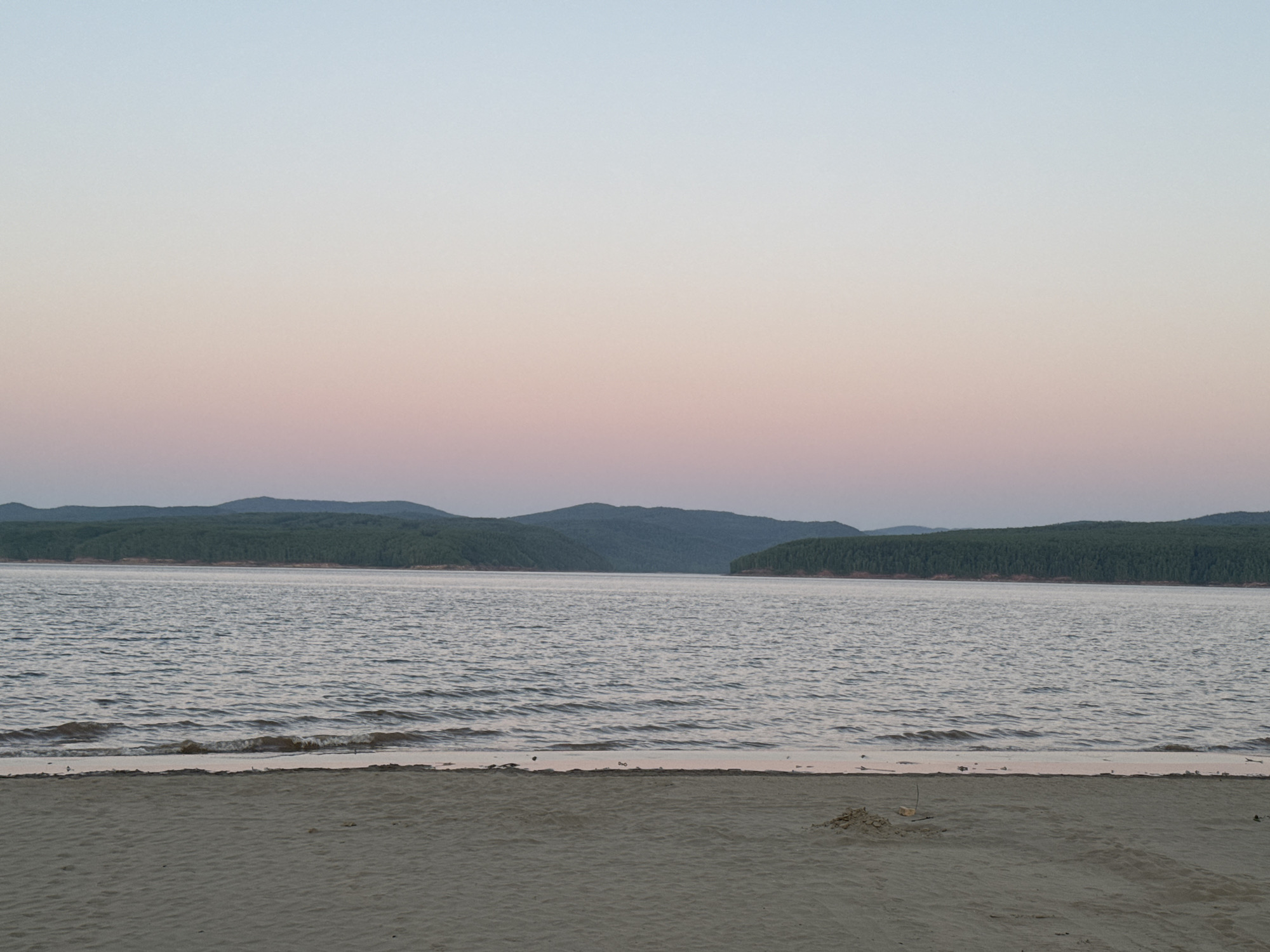 The image captures a serene landscape in the vicinity of Daurskoe, Russia, during twilight or dusk. The sky presents a soft gradient, transitioning from a pale blue at the top to light pink and lavender hues near the horizon line. A continuous chain of dark, densely forested hills or mountains is silhouetted against the colorful sky, forming the distant backdrop.
A wide body of water, likely a lake or reservoir, occupies the midground, its surface exhibiting gentle ripples and small waves that subtly reflect the muted sky colors. The water meets a sandy beach that spans the foreground of the image. The sand is light brown, showing faint texture and undulations. Near the waterline, a small, irregular pile of sand or pebbles is visible on the beach. No people, animals, or artificial structures are present within the scene, emphasizing a natural and undisturbed environment.