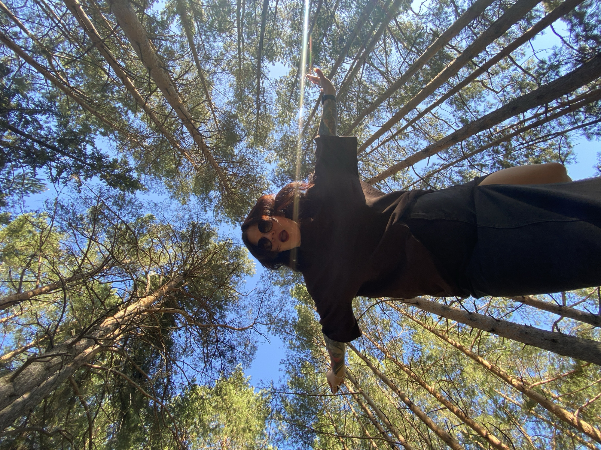 A low-angle shot looks upwards towards a person with red hair wearing sunglasses and dark clothing standing among tall trees in a forest. The person is reaching up with one arm, and a tattoo is visible on their forearm. The sky is visible through the trees. The location is Фоки, Russia.