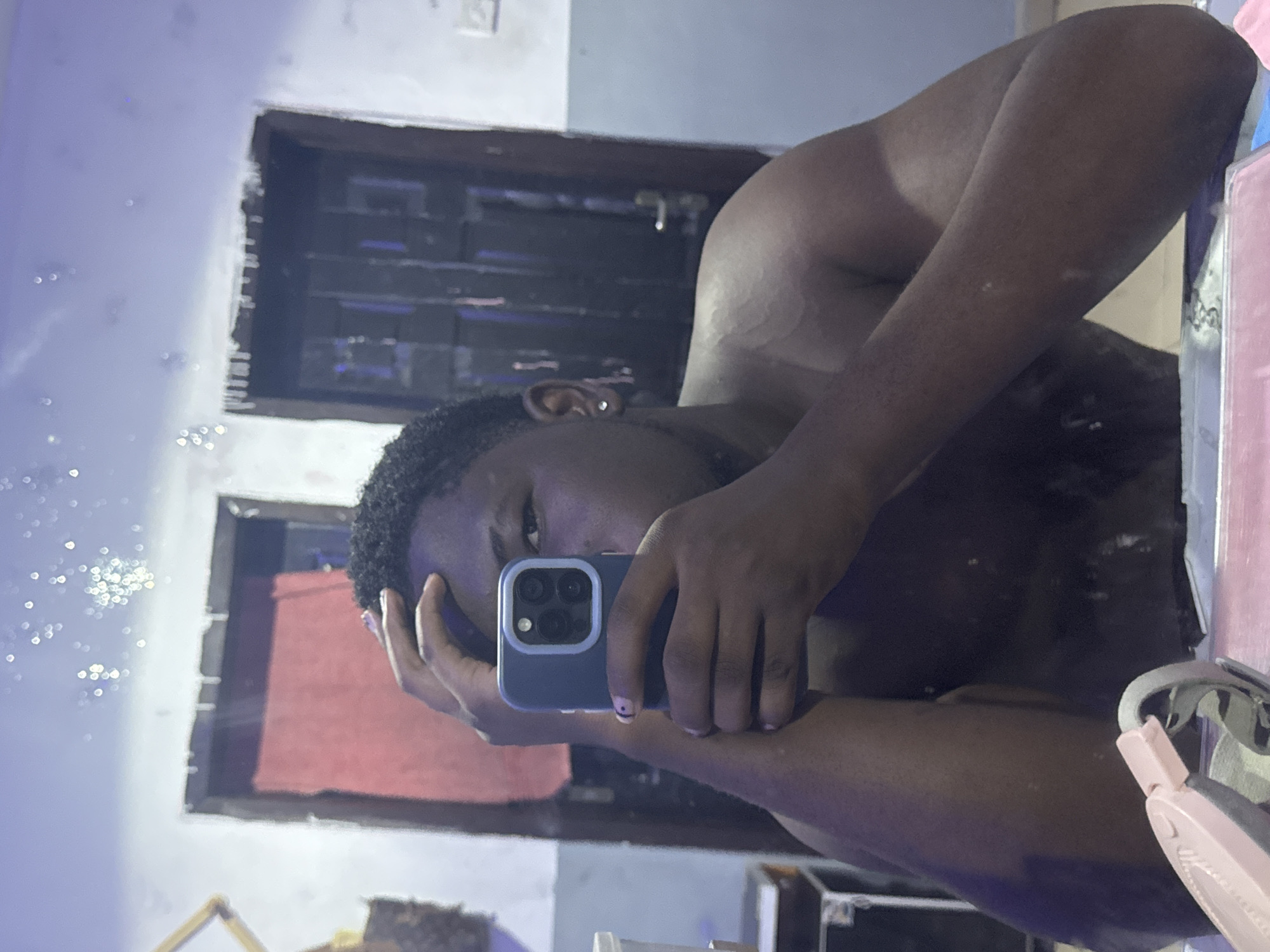 A dark-skinned individual with curly hair is taking a selfie in what appears to be a bathroom. The individual is holding a blue iPhone in front of their face, obscuring part of it. They have an earring in their left ear. The background includes a doorway with a dark interior and a red object visible inside. A mirror reflects the scene, and there are some light fixtures and other objects visible in the reflection. The location is given as Gbongan, Nigeria.