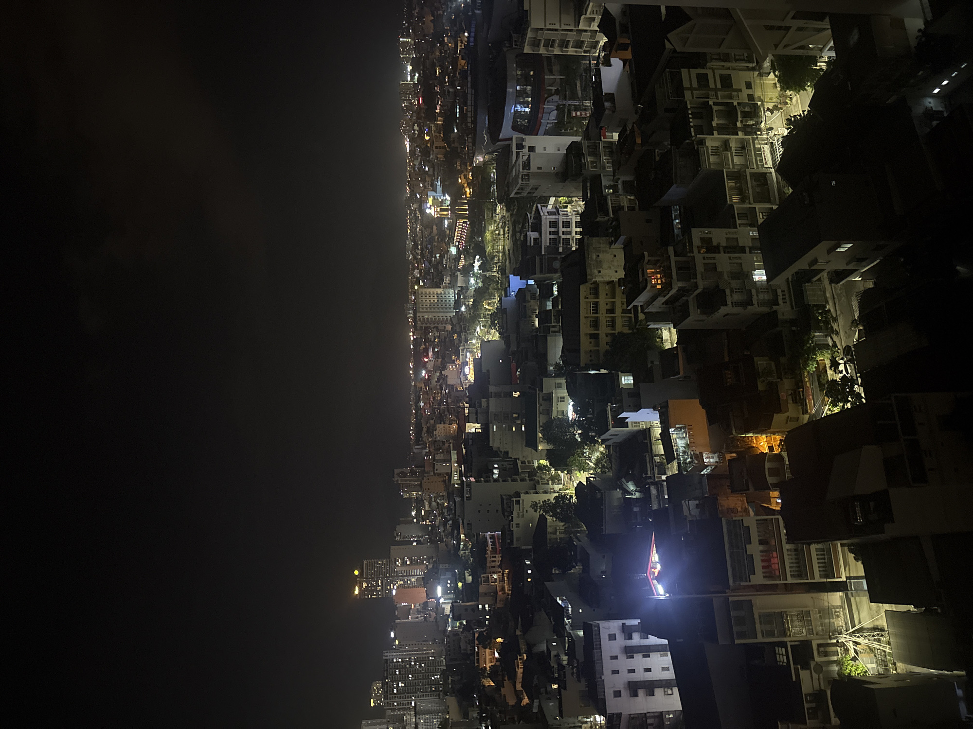 An aerial night view of the city of Nha Trang Ward, Vietnam, features a dense urban landscape illuminated by artificial lighting. The scene is split between a dark, overcast sky to the left and the illuminated city stretching along the right side. Buildings of varying heights are closely packed together, with the light from their windows and streetlights contributing to the overall glow of the cityscape. The absence of discernible people or specific actions directs the focus to the urban environment and its constructed elements. The photo orientation is rotated, so the city landscape runs vertically rather than horizontally.