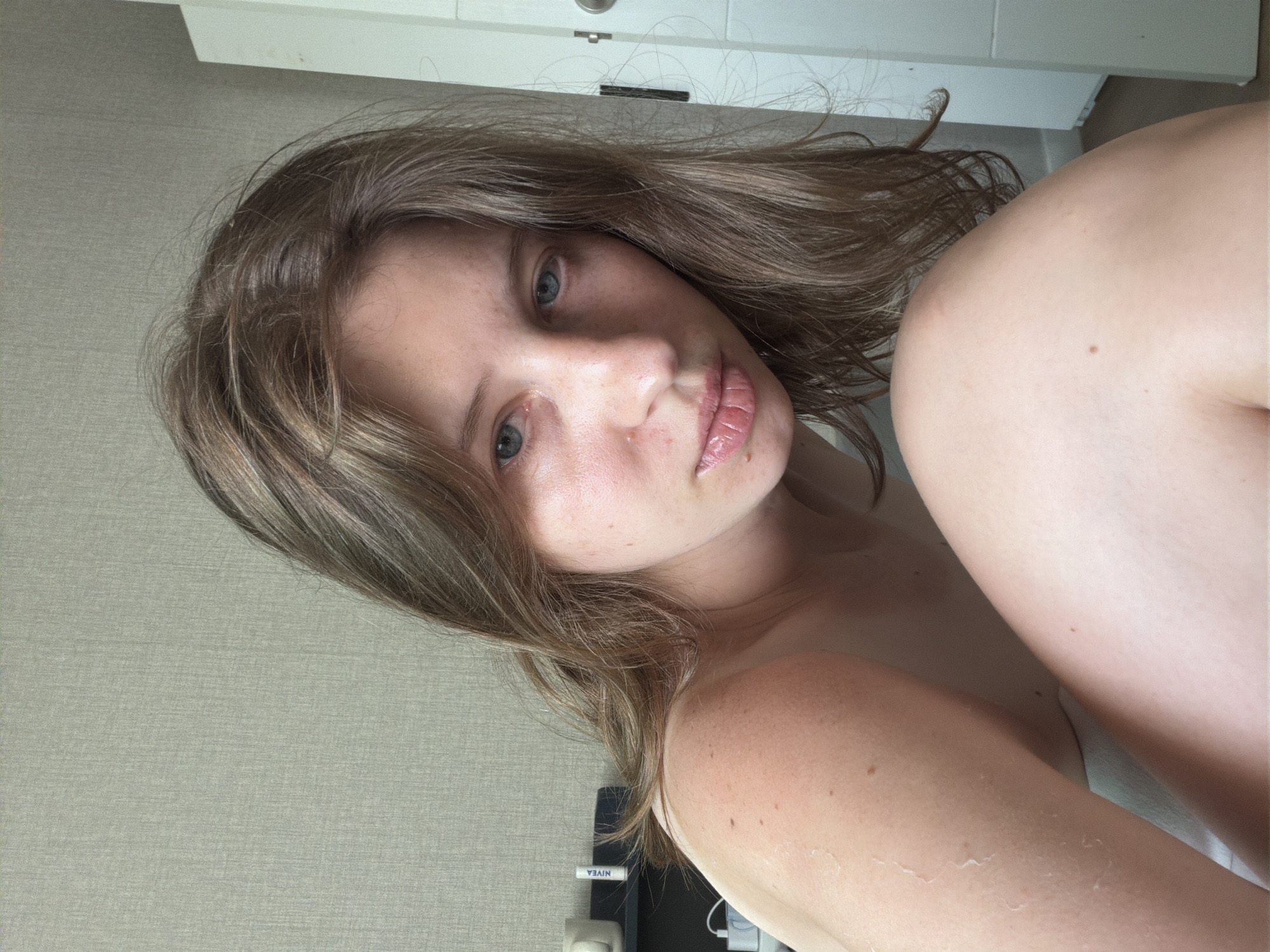 The image shows a close-up, head-and-shoulders portrait of a young woman with light brown hair and blue eyes. She is looking directly at the camera. Her skin appears slightly sunburnt on her shoulder and upper arm, with some flaking visible. Her lips are slightly parted and appear glossy. She is wearing a light-colored top that is off-the-shoulder. In the background, a textured wall is visible, along with a shelf holding a white mug and a tube of Nivea lip balm. A white door with a silver handle is also visible on the right side of the frame. The lighting suggests it is daytime. The location is not specified in the image.