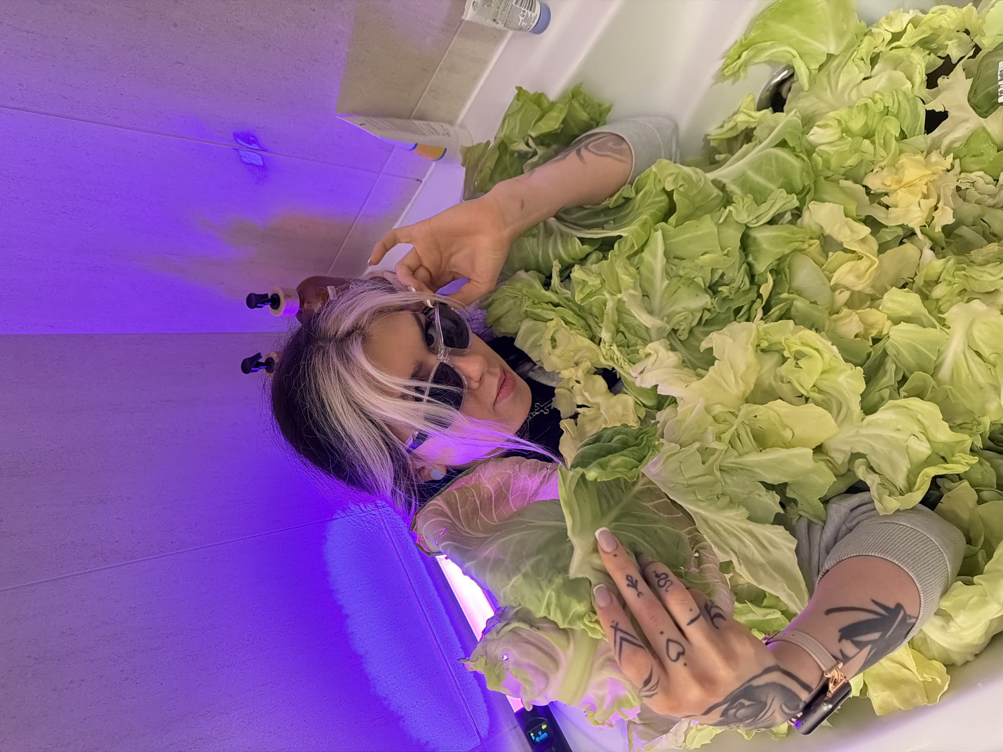 The image depicts an indoor bathroom scene with a single individual in a bathtub filled with numerous loose cabbage leaves, under prominent purple ambient lighting.
**Individual:** A female individual, centered in the frame, possesses long dark hair with distinct blonde/white highlights on the top and front. She wears dark sunglasses with clear frames and a light blue stud earring in her left ear. Her visible clothing includes a dark high-necked garment beneath a light grey long-sleeved top. Both arms are heavily tattooed with abstract and geometric designs. Her left hand, featuring manicured nails, displays small tattoos including a heart and an arrow-like symbol, and holds a large cabbage leaf. A dark-faced smartwatch with a light-colored band is worn on her left wrist. Her right hand is positioned near her sunglasses. The individual is looking directly towards the camera.
**Objects and Environment:** The individual is seated in a white bathtub, which is largely filled with fresh, light green cabbage leaves. The surrounding bathroom walls are tiled in a light color, with visible grout lines. A strong purple light emanates from an unseen source, likely an LED strip, illuminating the upper wall sections and casting reflections. A device with a digital display and a purple light source is visible on the left side of the frame. On the right rim of the bathtub, two pump bottles (one brown, one clear/amber) and two plastic containers (one white tube, one clear bottle with a blue cap) are present. A partial product label on a cabbage leaf near the tub's drain reads "VOLI. Budva."
**Actions and Interactions:** The individual is posing within the cabbage-filled bathtub, holding a cabbage leaf, and appearing to adjust her sunglasses. There are no other individuals present or interactions depicted.
**Location:** The scene is an indoor bathroom. The presence of a "VOLI. Budva" label on a cabbage leaf indicates that the produce was sourced from a Voli supermarket in Budva, Montenegro. This links the scene contextually to the city of Budva; however, the specific address or type of establishment (e.g., private residence, hotel) for the bathroom itself within Budva is not discernible from the image.