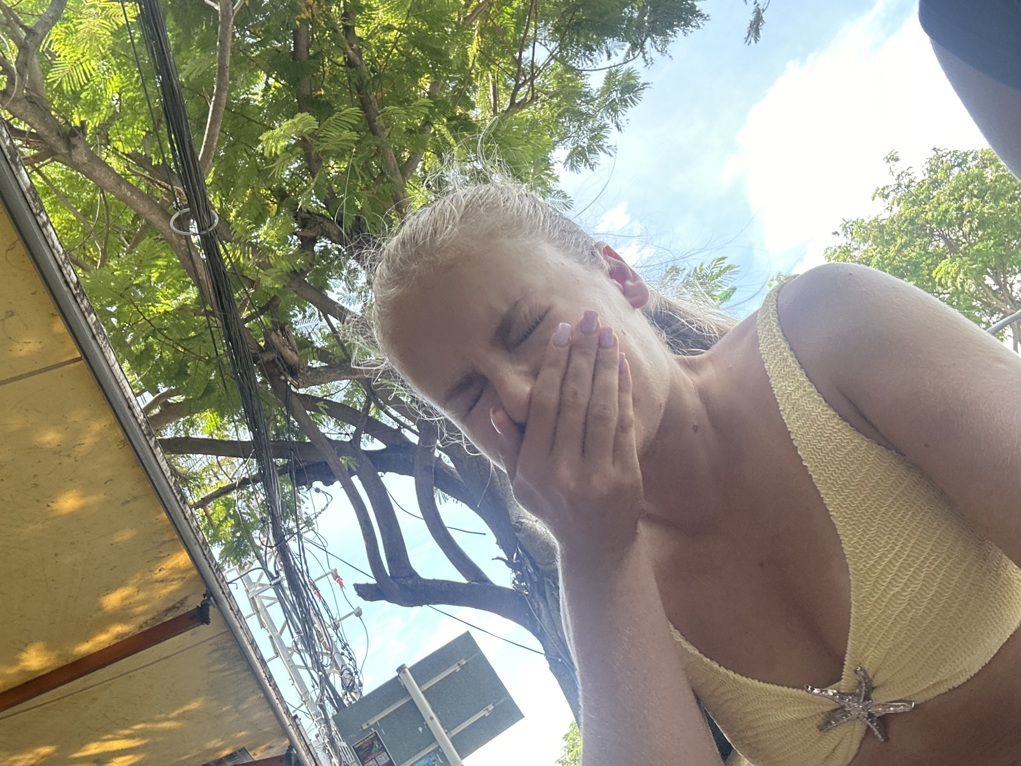 A young Caucasian woman with blonde hair is outdoors, holding her hand over her mouth. She is wearing a yellow bikini top adorned with a starfish. The background consists of lush green trees, a bright blue sky with white clouds, and a yellow awning. Electrical wires and a street sign are visible in the upper left portion of the image. The scene suggests a sunny day, possibly in a tropical location. The image is oriented with the top of the woman's head towards the bottom.