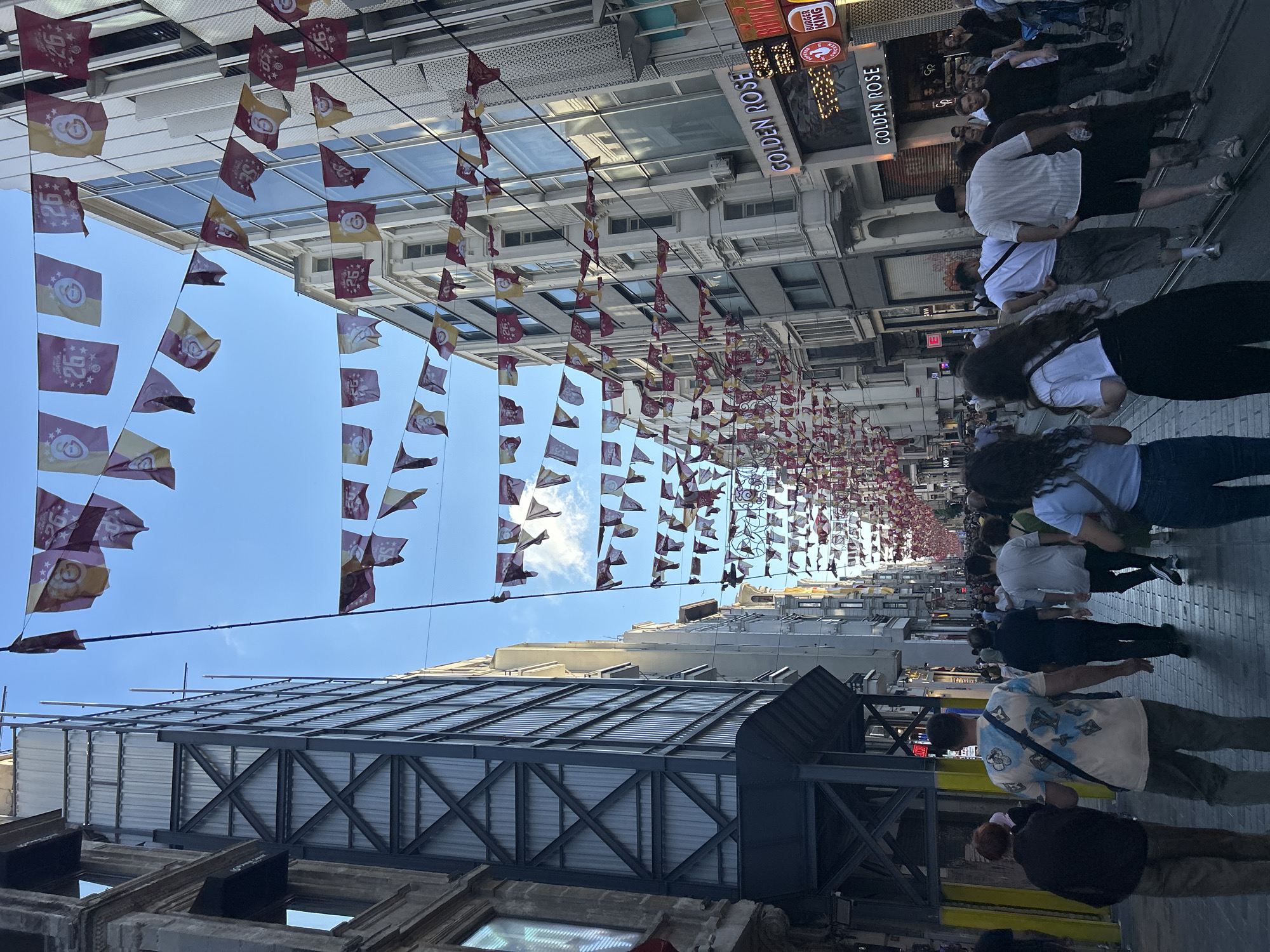 The image shows a crowded pedestrian street in Beyoğlu, Turkey, likely Istiklal Avenue. A long string of decorative flags, predominantly red and yellow with a crest and the number "26" on them, are strung across the street, creating a festive atmosphere. Buildings line both sides of the street, with some appearing to be under construction or renovation, indicated by scaffolding and covered facades. Several people are walking in both directions on the cobblestone street, which also has tram tracks embedded in it. Stores and businesses, including "GOLDEN ROSE" and "BURGER KING," are visible with their signage. The sky is blue with some clouds, and the overall lighting suggests it is daytime. The people are dressed in casual summer attire.