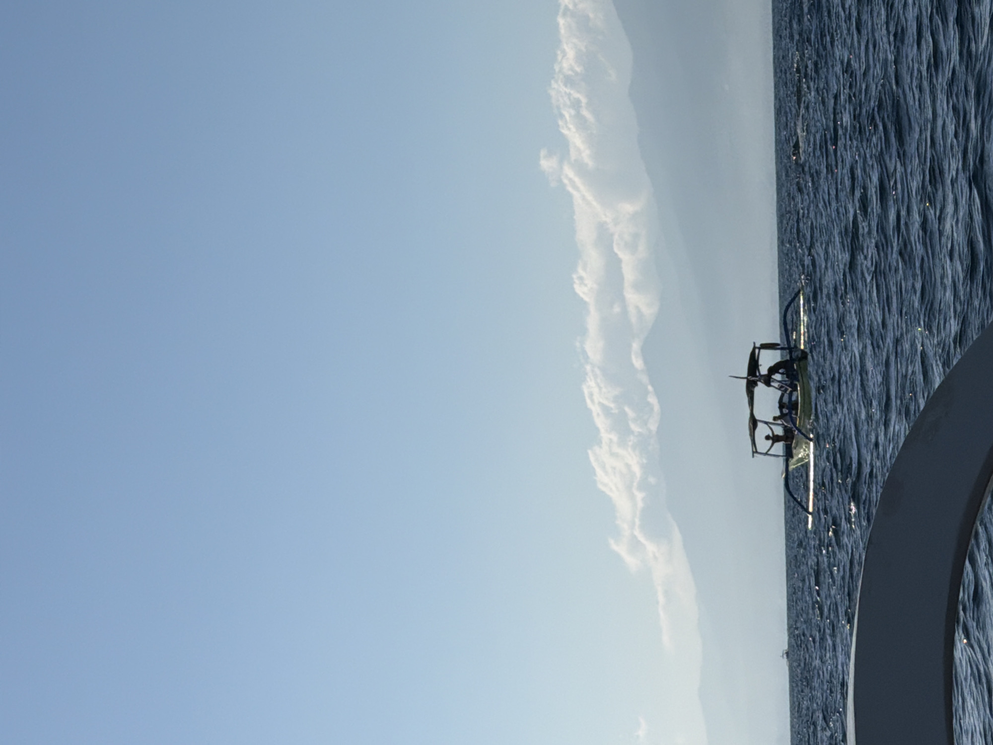 An eye-level, vertical medium shot captures a wide expanse of choppy, dark blue ocean under a clear, pale blue sky. In the mid-ground, a traditional Balinese outrigger boat, painted yellow with blue outriggers, sails across the water. The boat has a dark canopy and is occupied by at least four people, one of whom stands with arms raised. In the distance, a hazy mountain range rises from the horizon, partially obscured by a thin layer of white clouds. Another, smaller boat is visible in the far distance on the left side of the frame. The foreground features the curved edge of another boat, a dark grey object that occupies the bottom left quadrant of the image. The overall scene depicts a serene day at sea with clear visibility and calm weather.