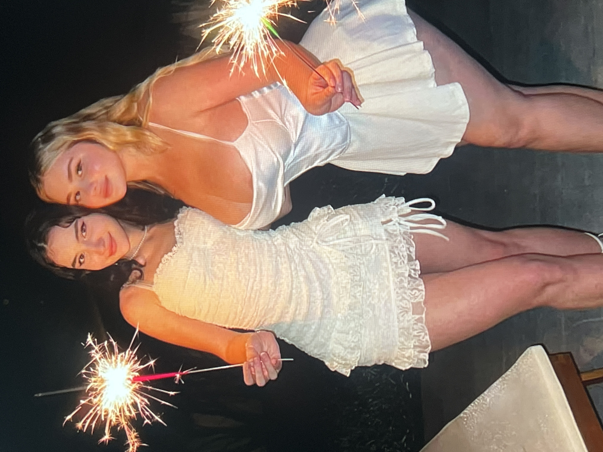 Two young women are holding sparklers in the dark. The woman in the foreground has dark hair and is wearing a white, textured dress with lace trim. She is looking at the camera with a slight smile. The woman in the background has blonde hair and is wearing a white slip dress. She is also looking towards the camera and smiling. Both women are holding sparklers that are lit and emitting sparks. The background is dark, suggesting it is nighttime. The overall mood appears festive and celebratory. The text "Vanadzor, Armenia" is visible in the prompt.